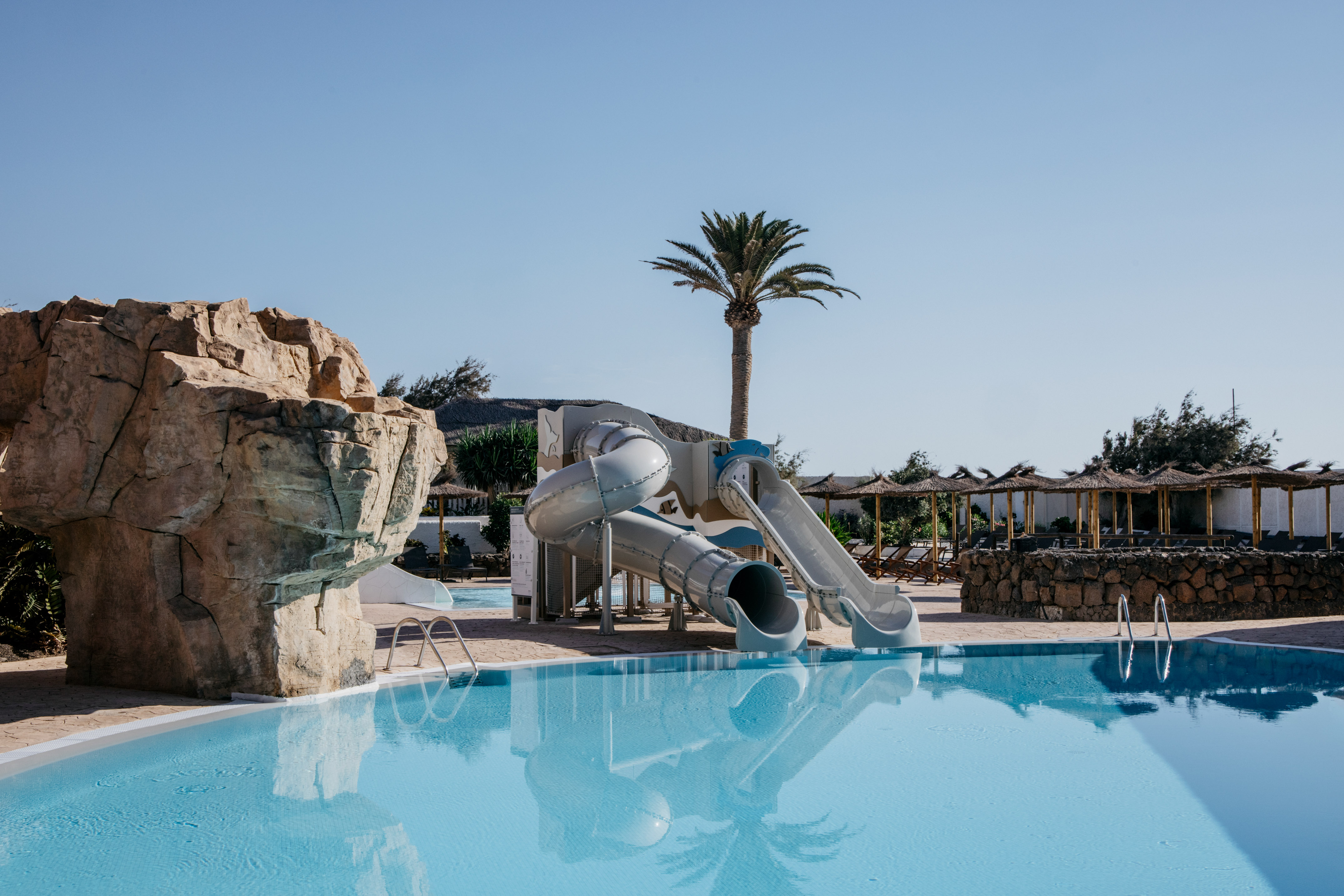 Resort swimming pool with water slides, palm tree, and thatched parasols.