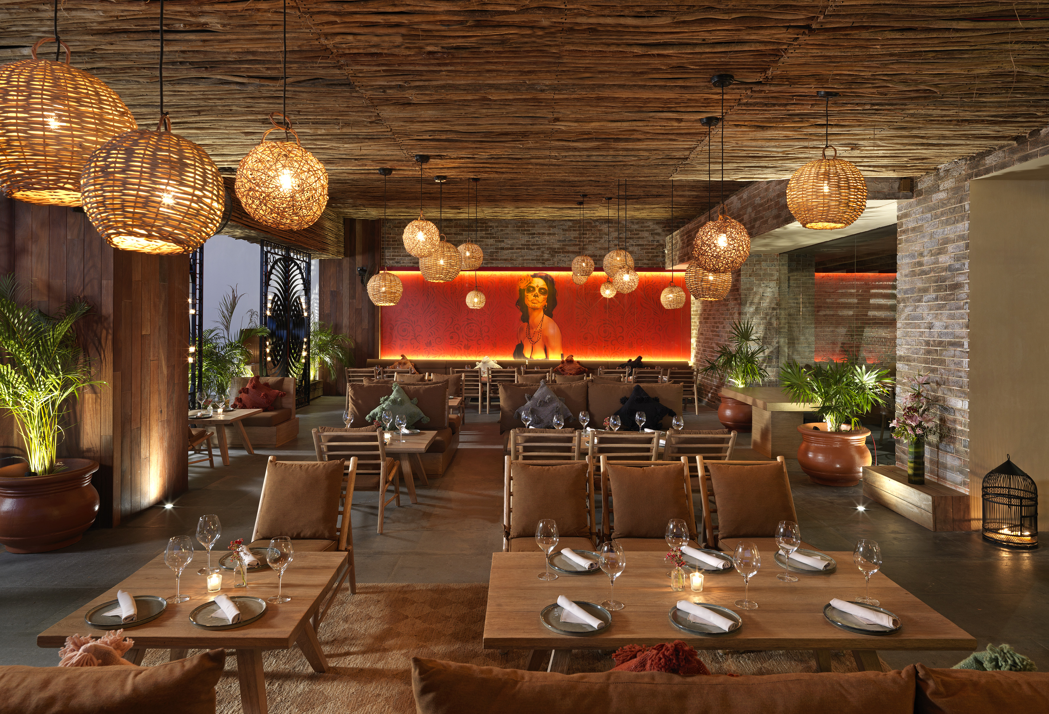 Lush restaurant interior with woven lights, wooden tables, tropical plants, and a vibrant mural.