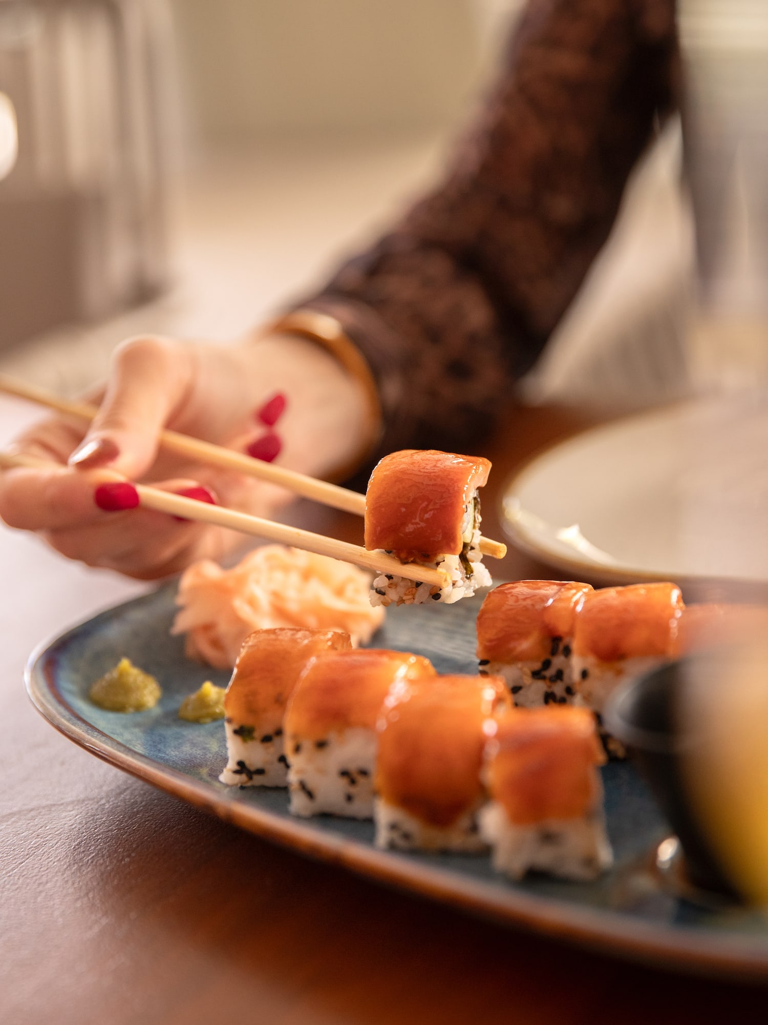 Woman's hand with red nails lifts a salmon sushi roll with chopsticks.