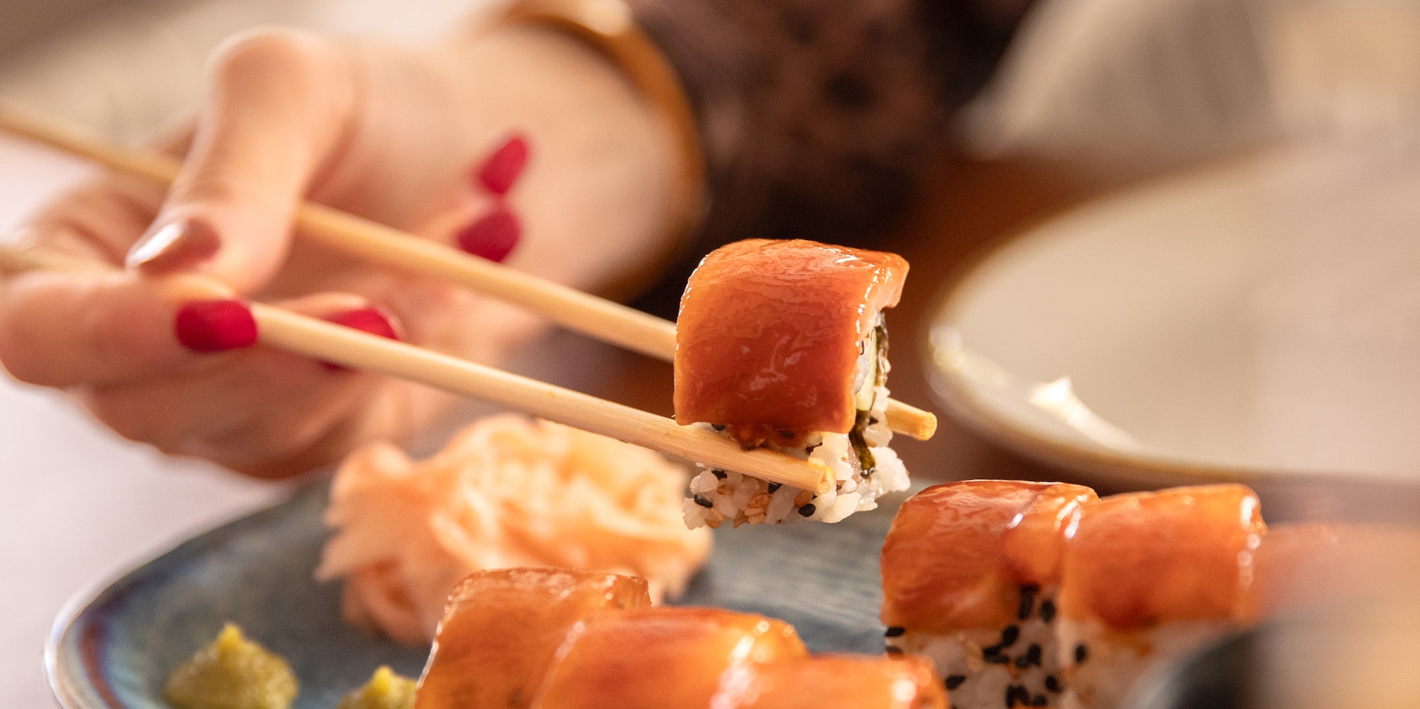 Hand with chopsticks lifts sushi roll from a plate with maki, ginger, wasabi.