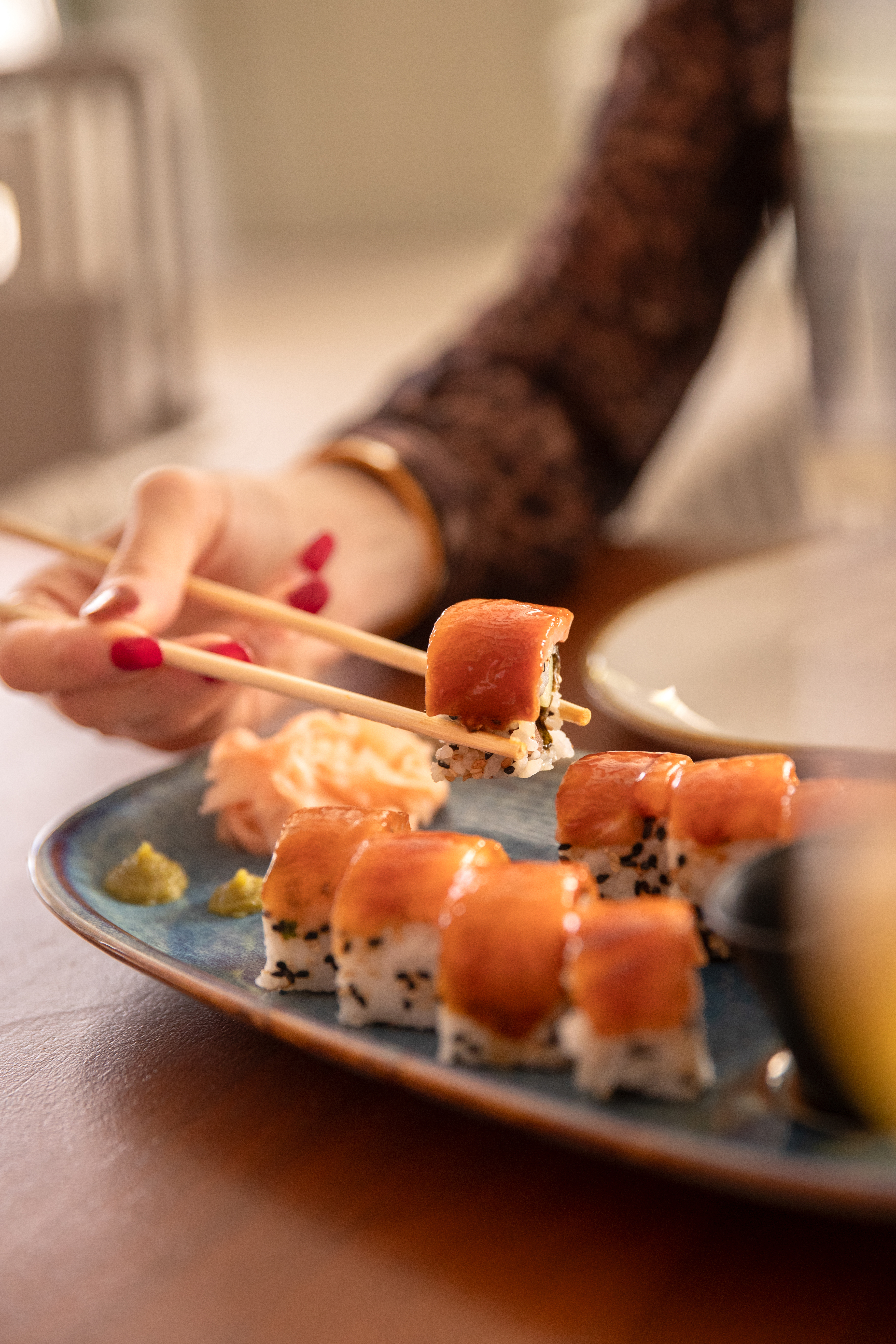 Woman's hand with red nails lifts a salmon sushi roll with chopsticks.