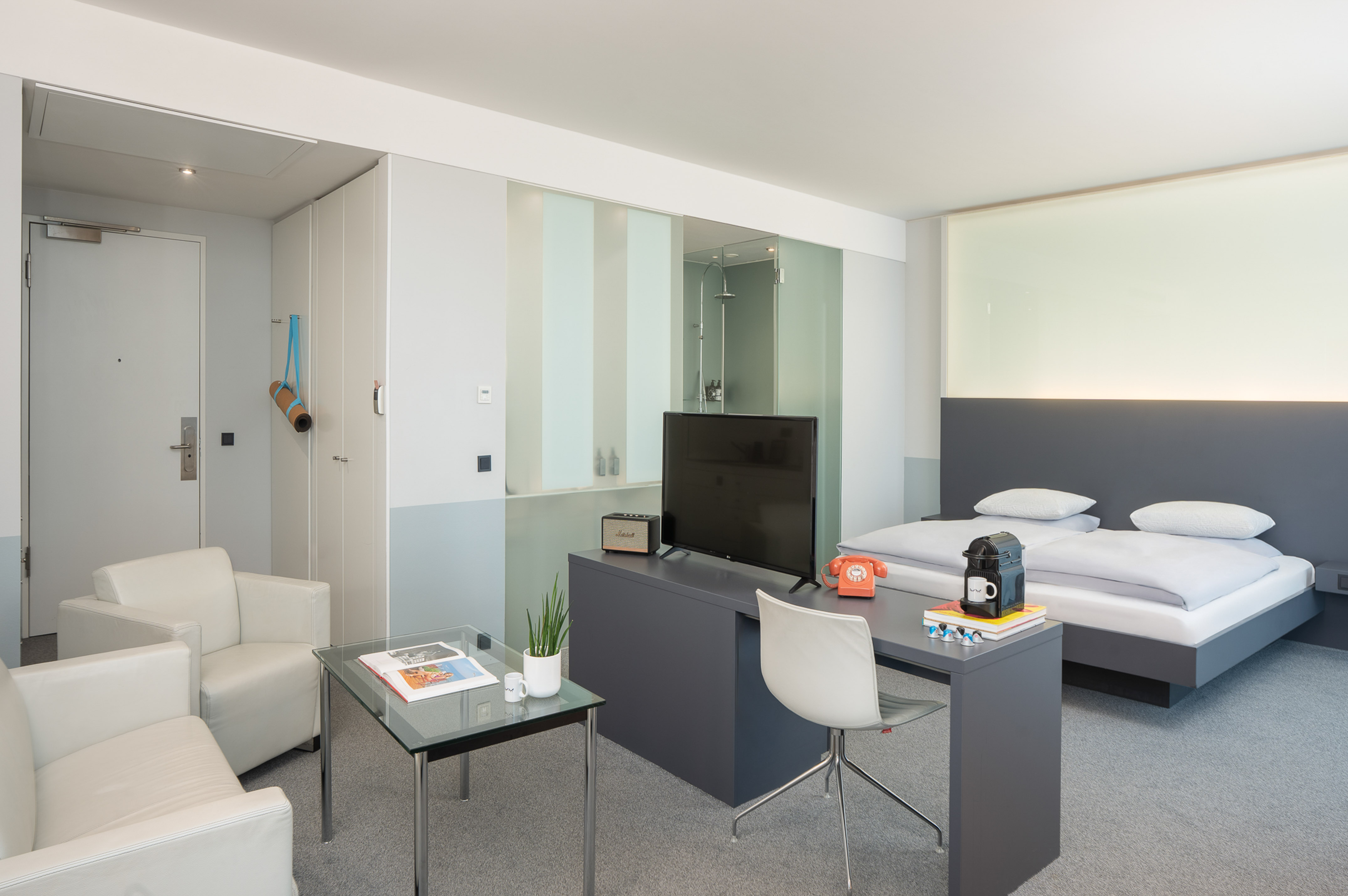 Modern hotel room with bed, desk, TV, white armchairs, and glass shower.