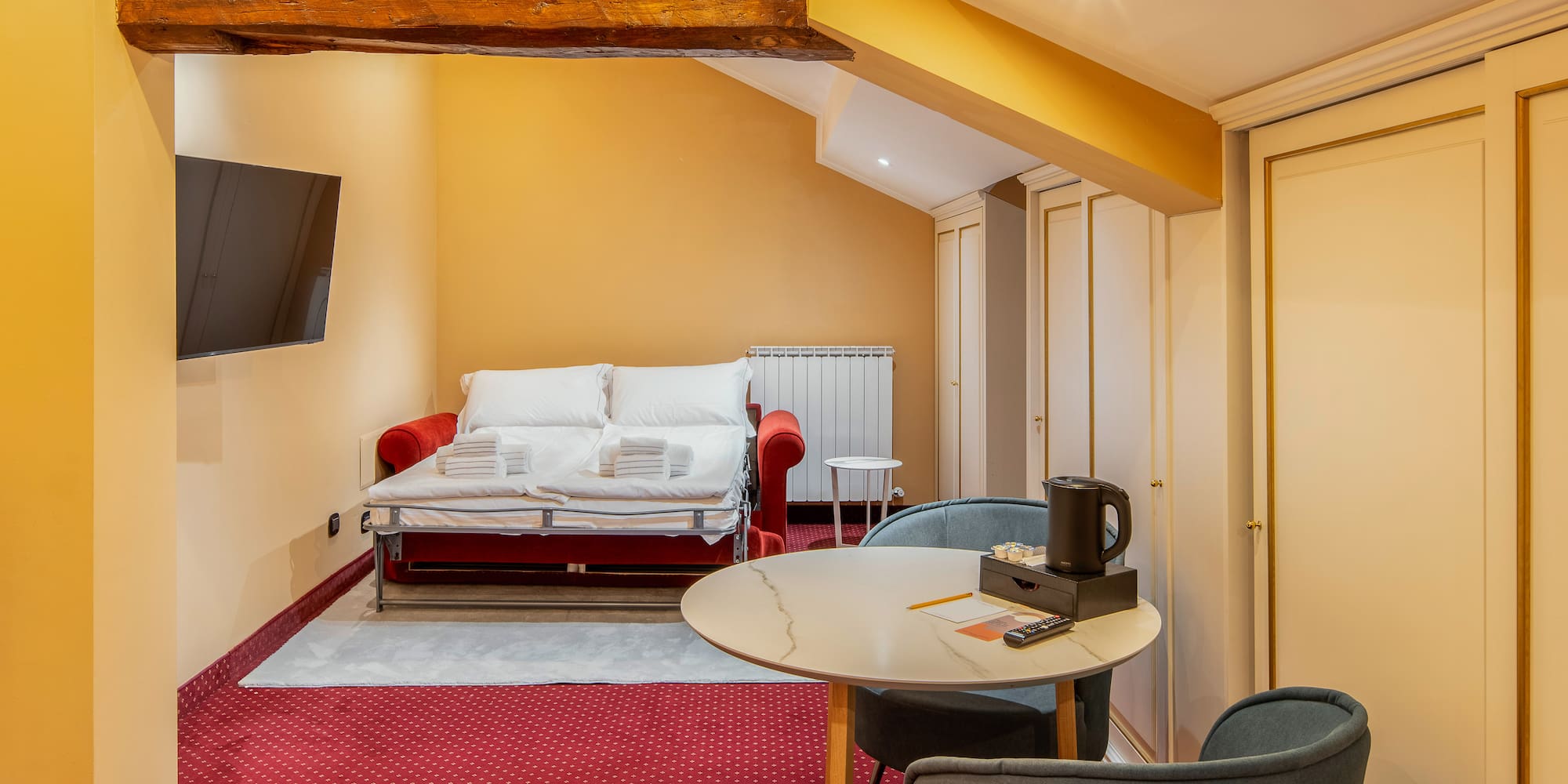Cozy hotel studio with red sofa bed, TV, dining table, and exposed wooden beam.