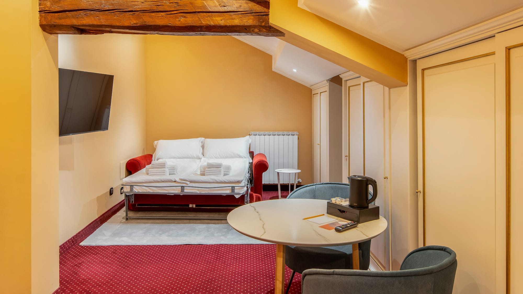 Cozy hotel studio with red sofa bed, TV, dining table, and exposed wooden beam.