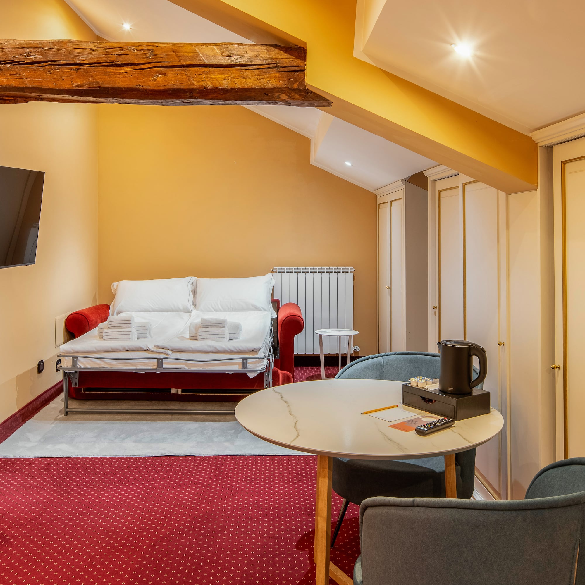 Cozy hotel studio with red sofa bed, TV, dining table, and exposed wooden beam.