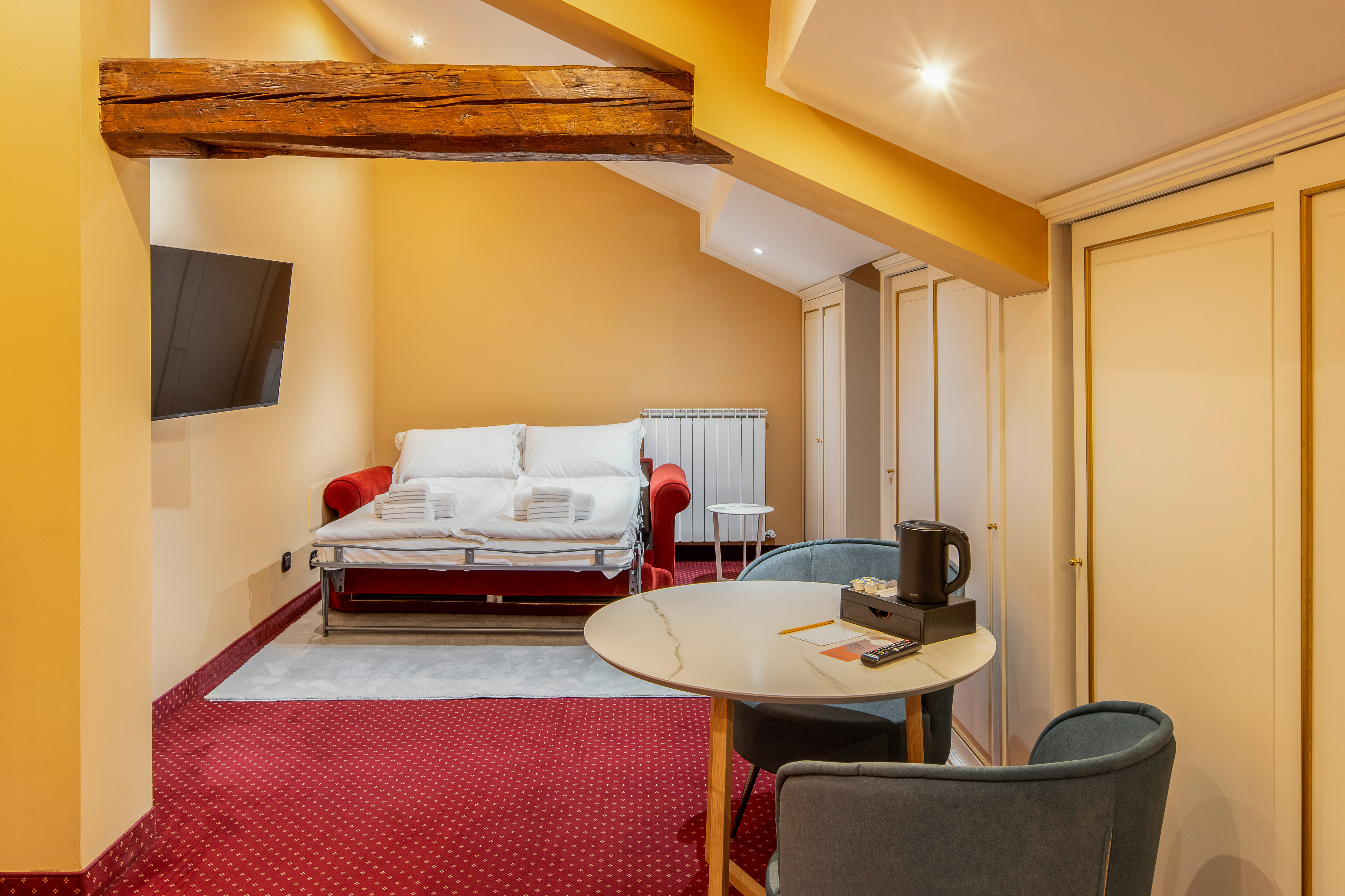 Cozy hotel studio with red sofa bed, TV, dining table, and exposed wooden beam.