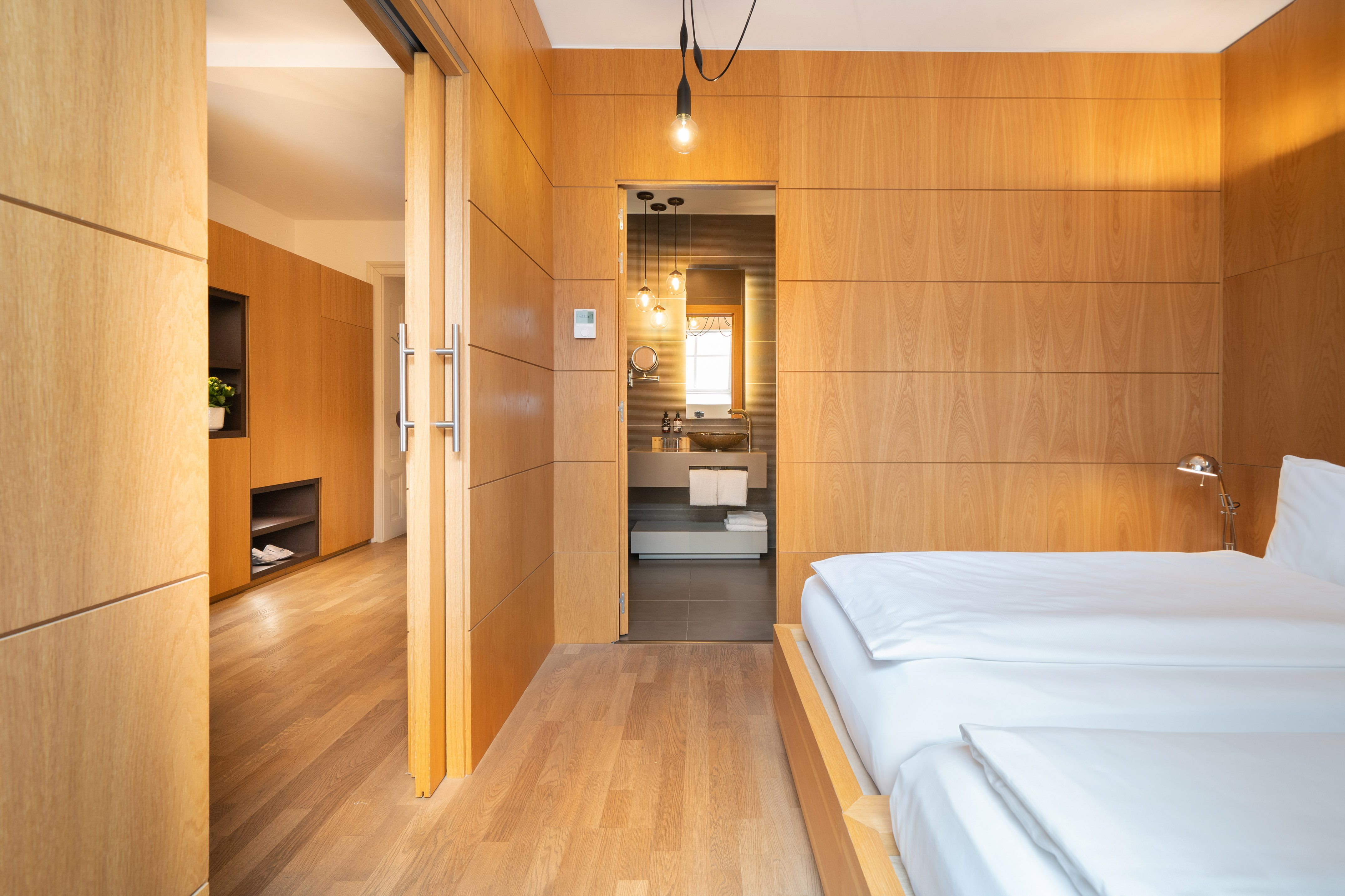 Modern hotel bedroom with wood paneling, white bed, and open en-suite bathroom.