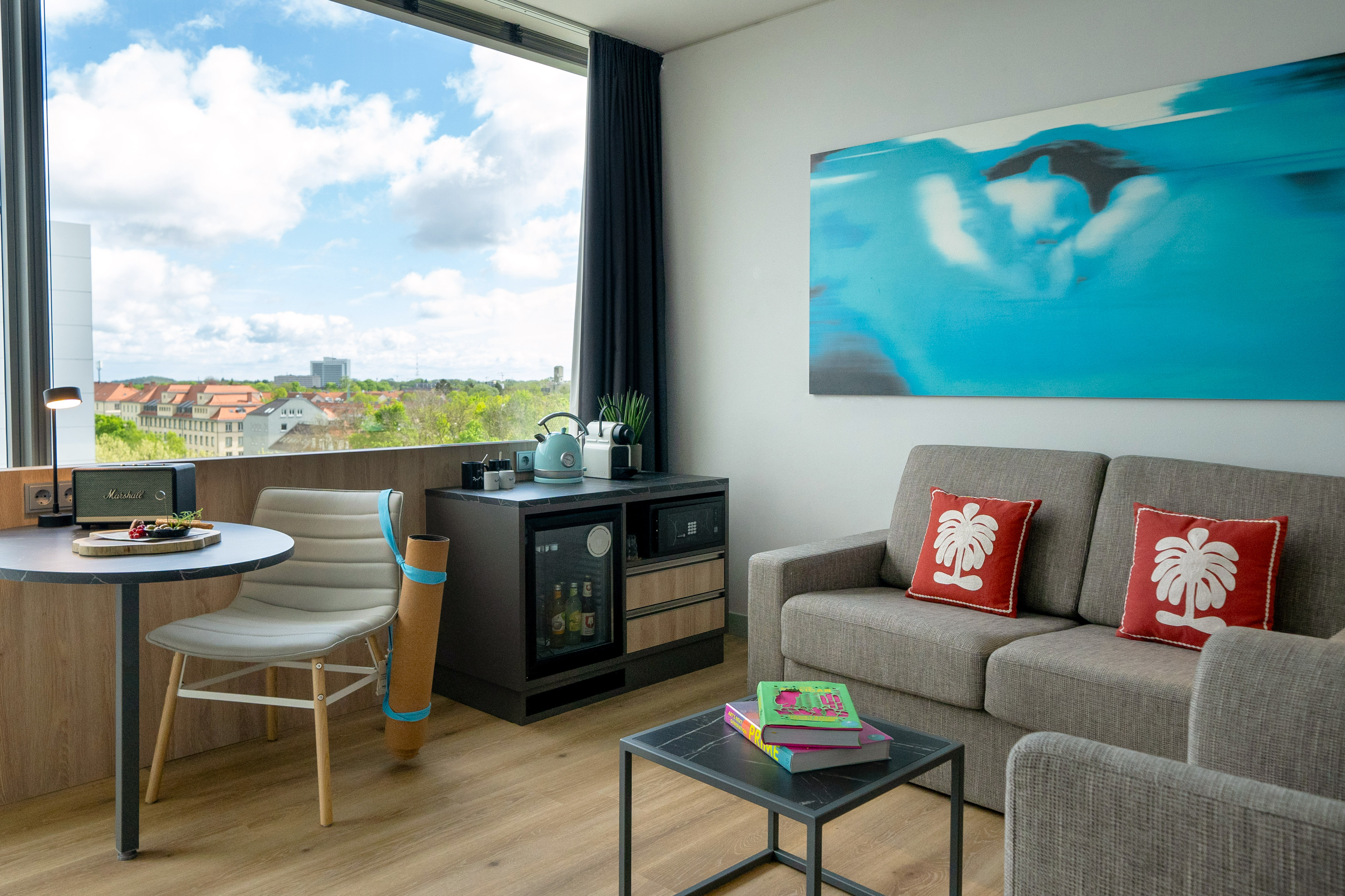 Bright modern hotel room with city view, sofa, desk, and minibar.