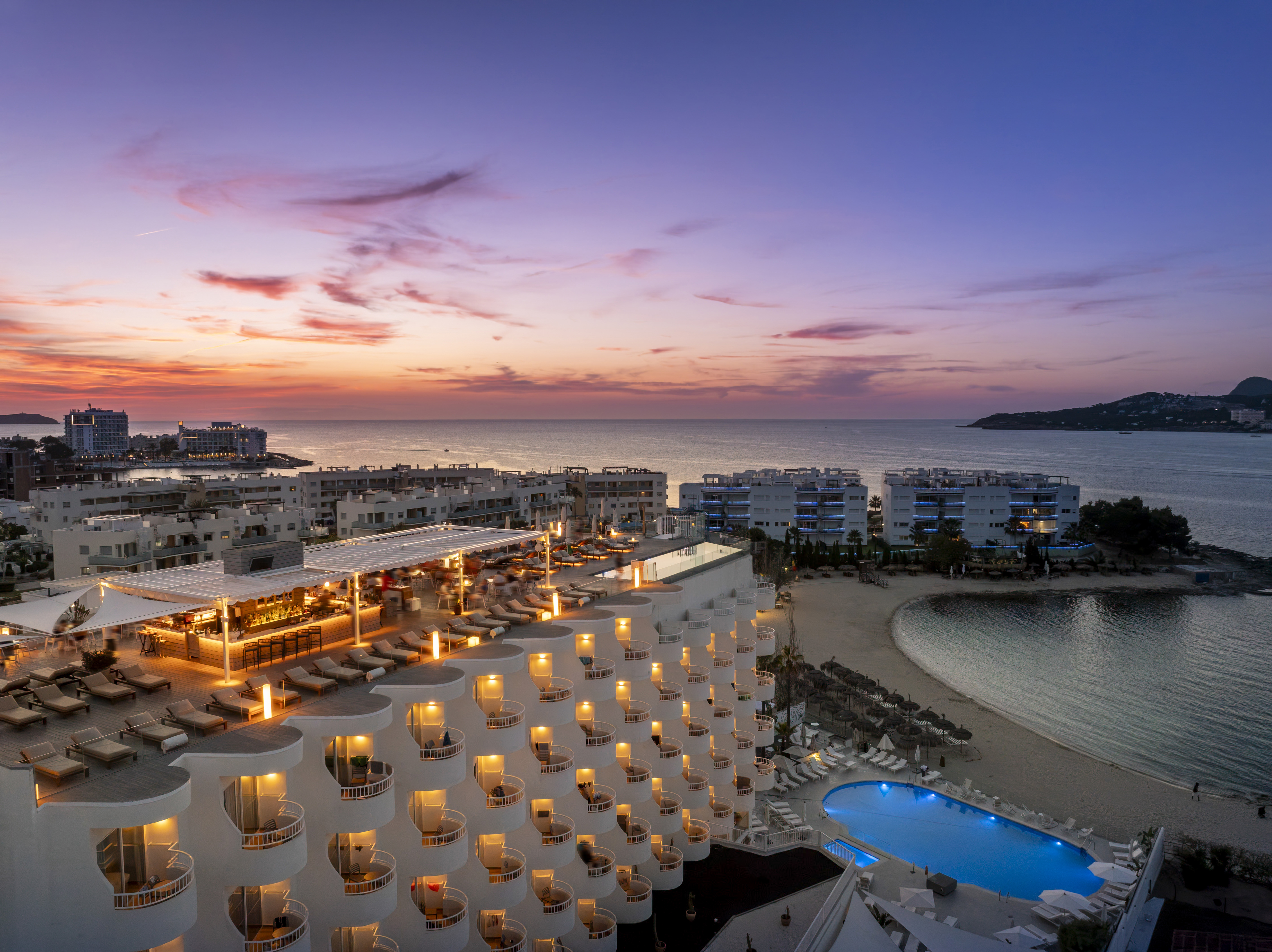 Lit coastal hotel, rooftop bar, pool, and beach at sunset.