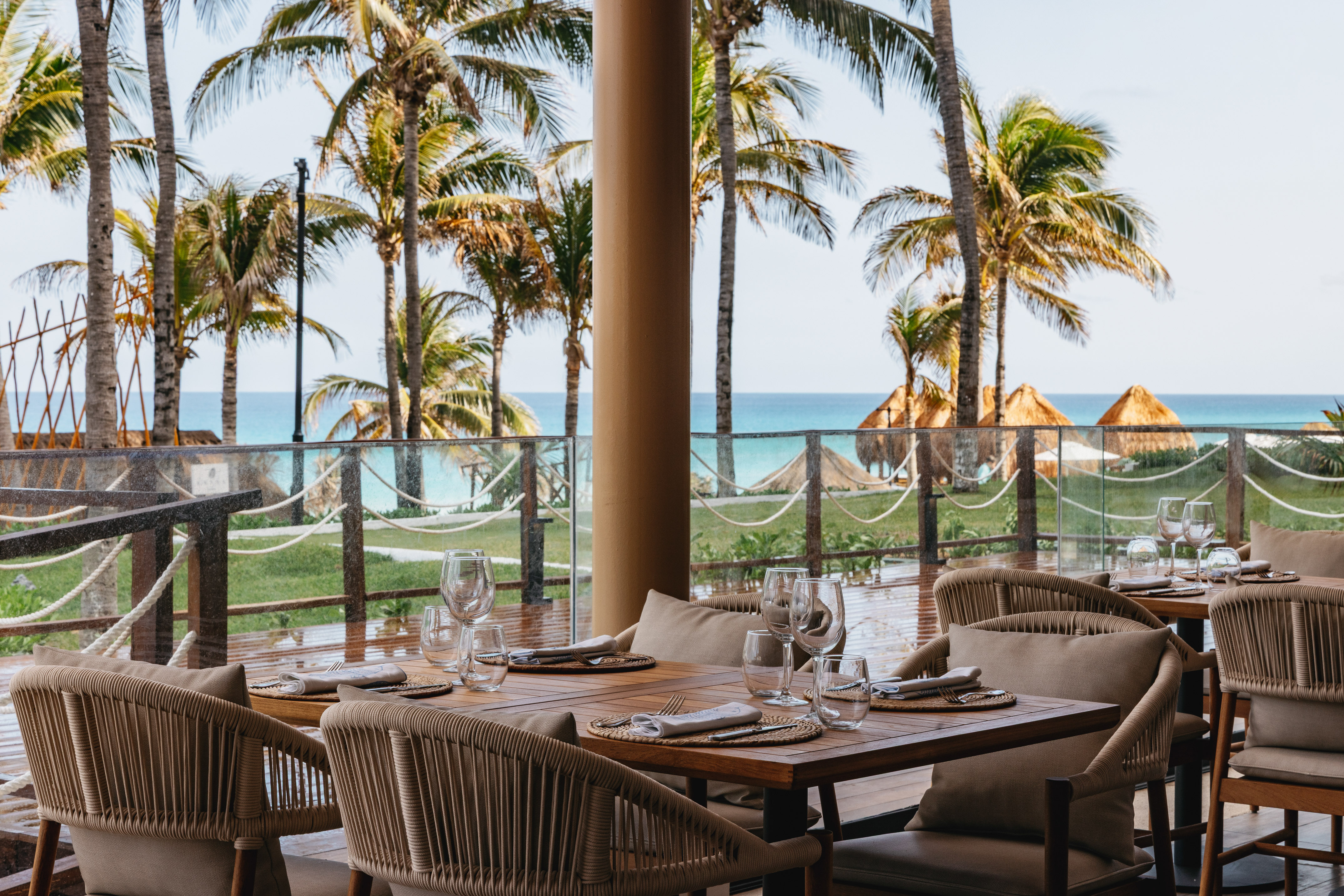 Beachfront restaurant dining area overlooking ocean, palm trees, and palapas.