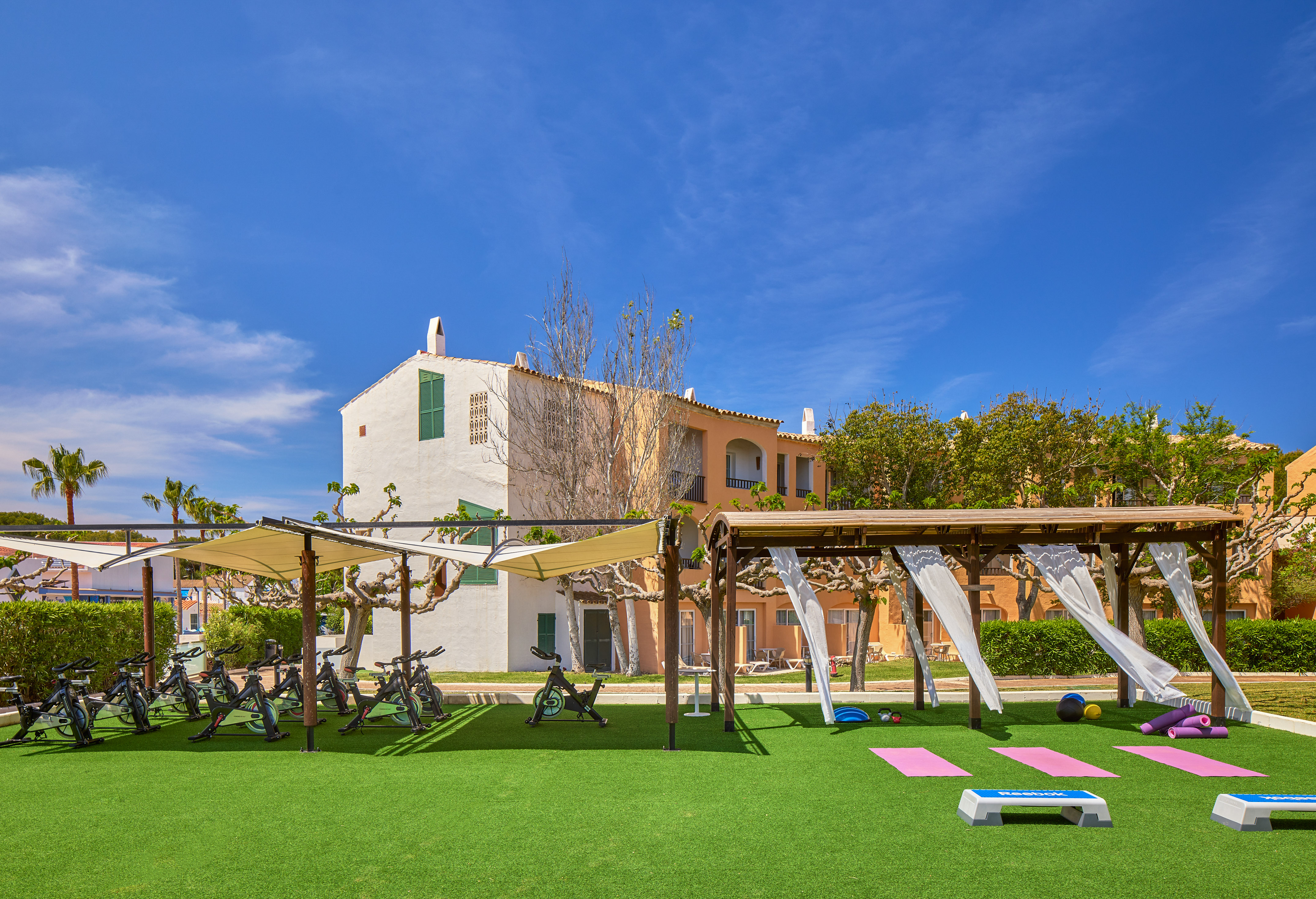 Outdoor resort gym: spinning bikes, yoga mats, and fitness equipment on turf.