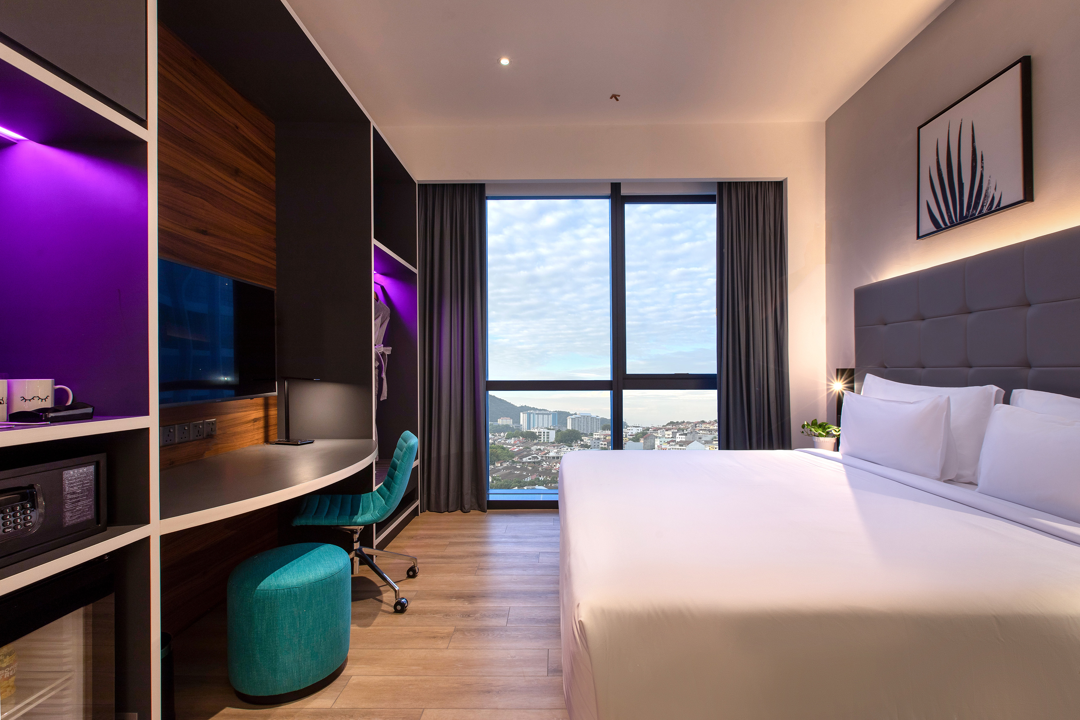 Modern hotel room with bed, work desk, city view, and purple LED lighting.