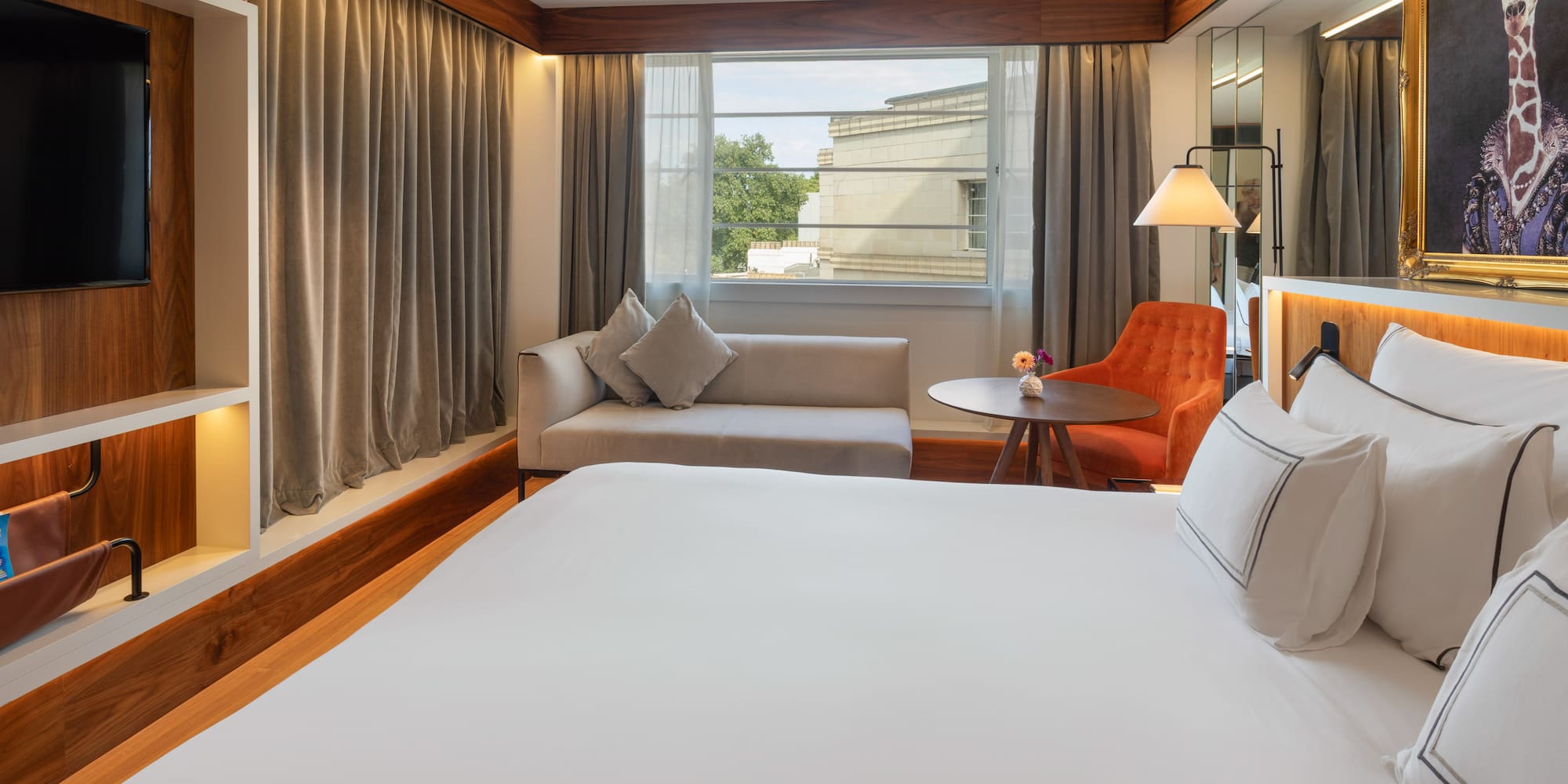 Modern hotel room with large bed, sofa, window view, and giraffe artwork.