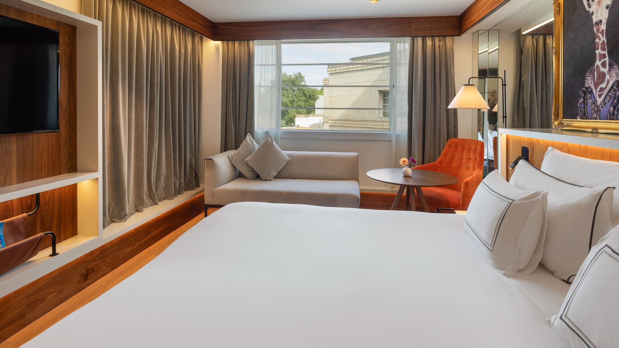 Modern hotel room with king bed, sofa, orange armchair, and giraffe art.