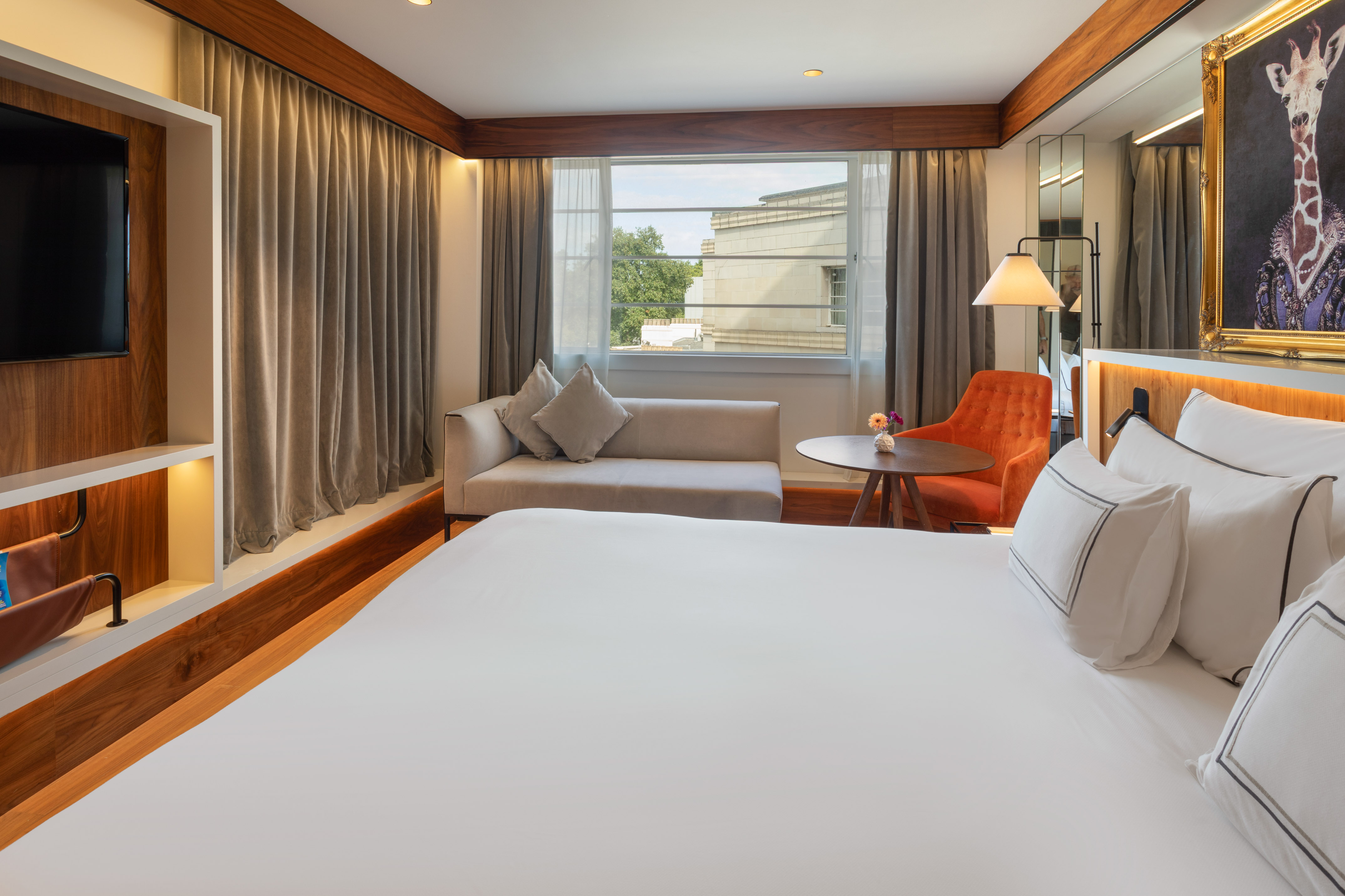 Modern hotel room with king bed, sofa, orange armchair, and giraffe art.