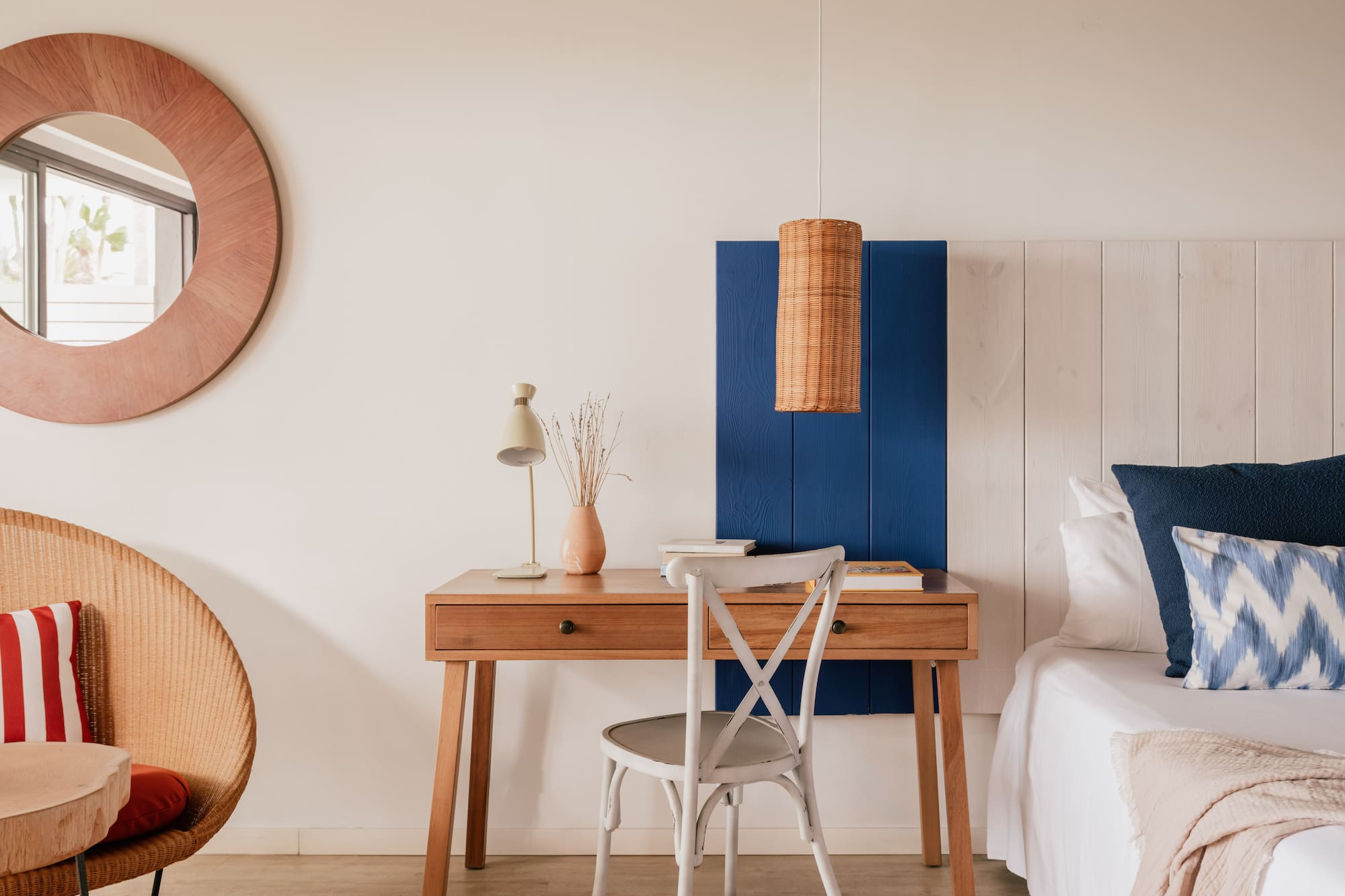 Habitación de hotel con escritorio de madera, cama blanca, espejo y detalles azul y mimbre.