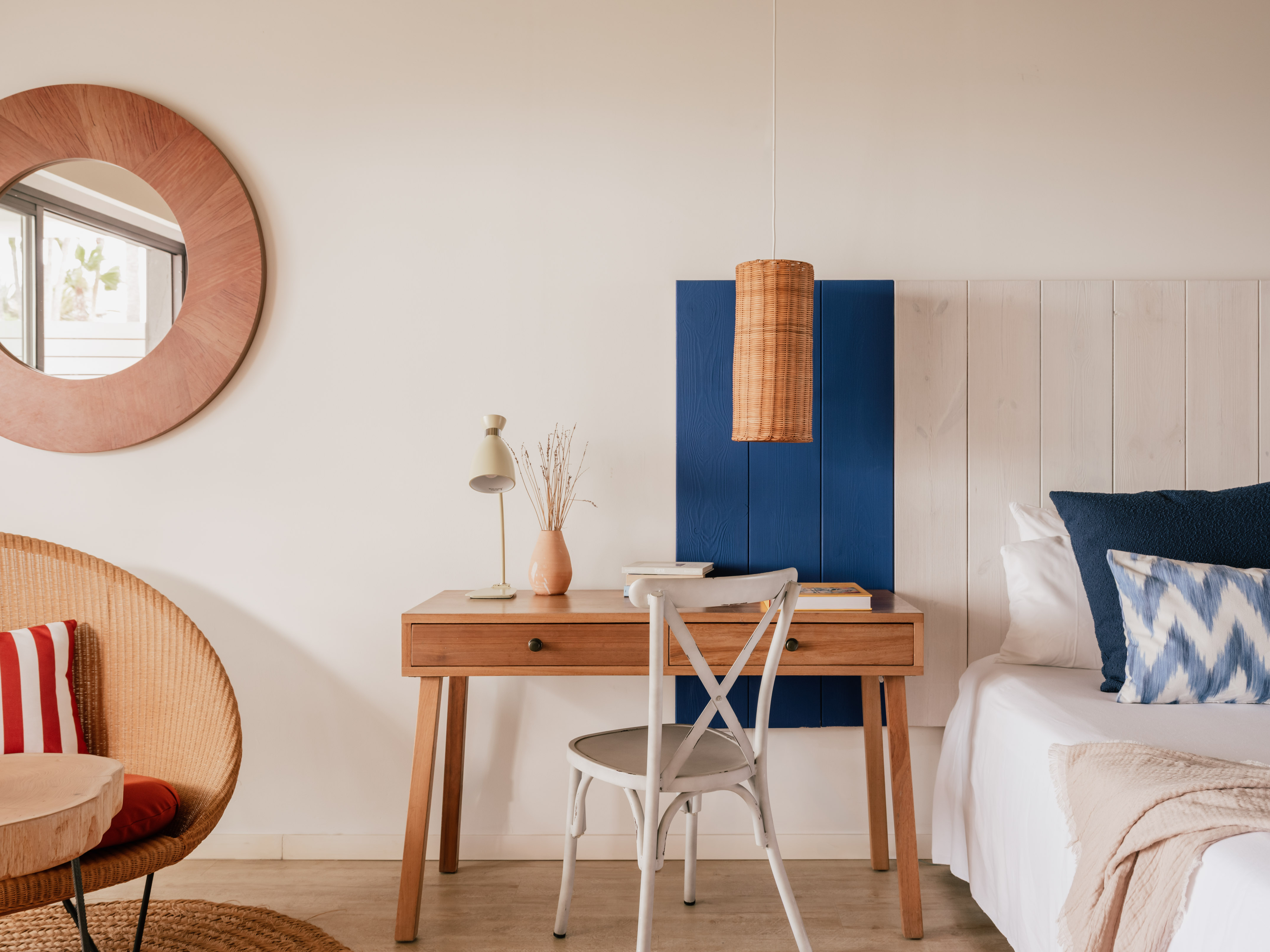 Habitación de hotel con escritorio de madera, cama blanca, espejo y detalles azul y mimbre.