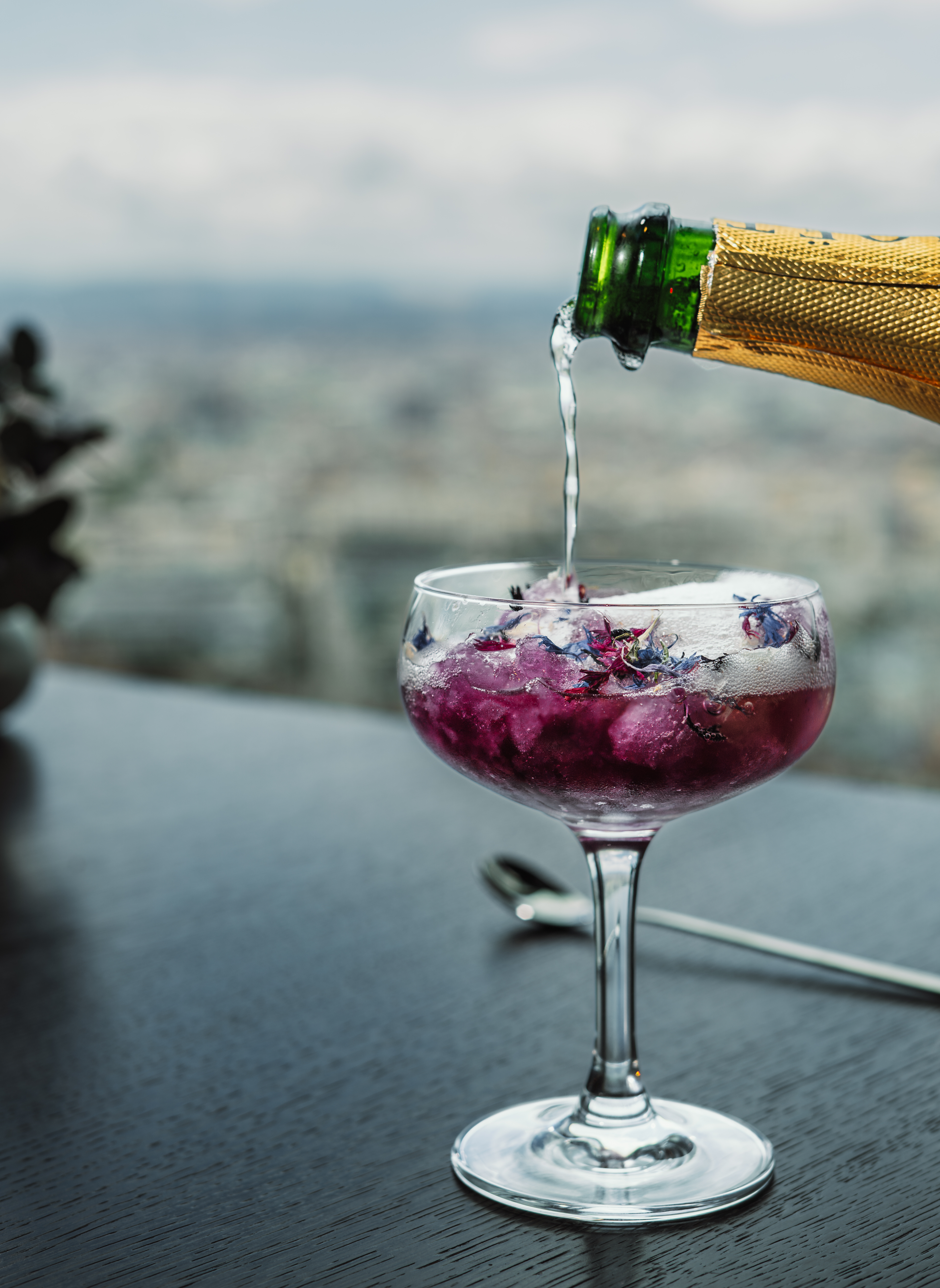 Champagne poured into coupe glass with purple floral cocktail and city view.