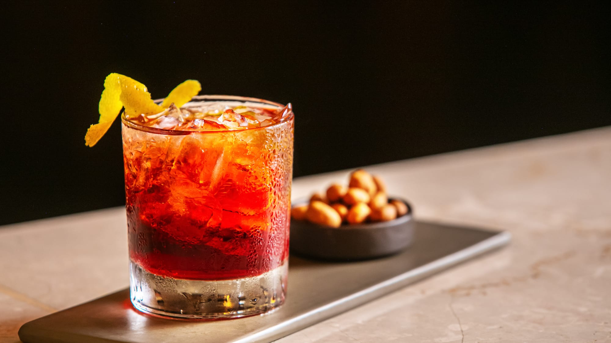Red cocktail with lemon peel, ice, and peanuts on a tray at a bar.