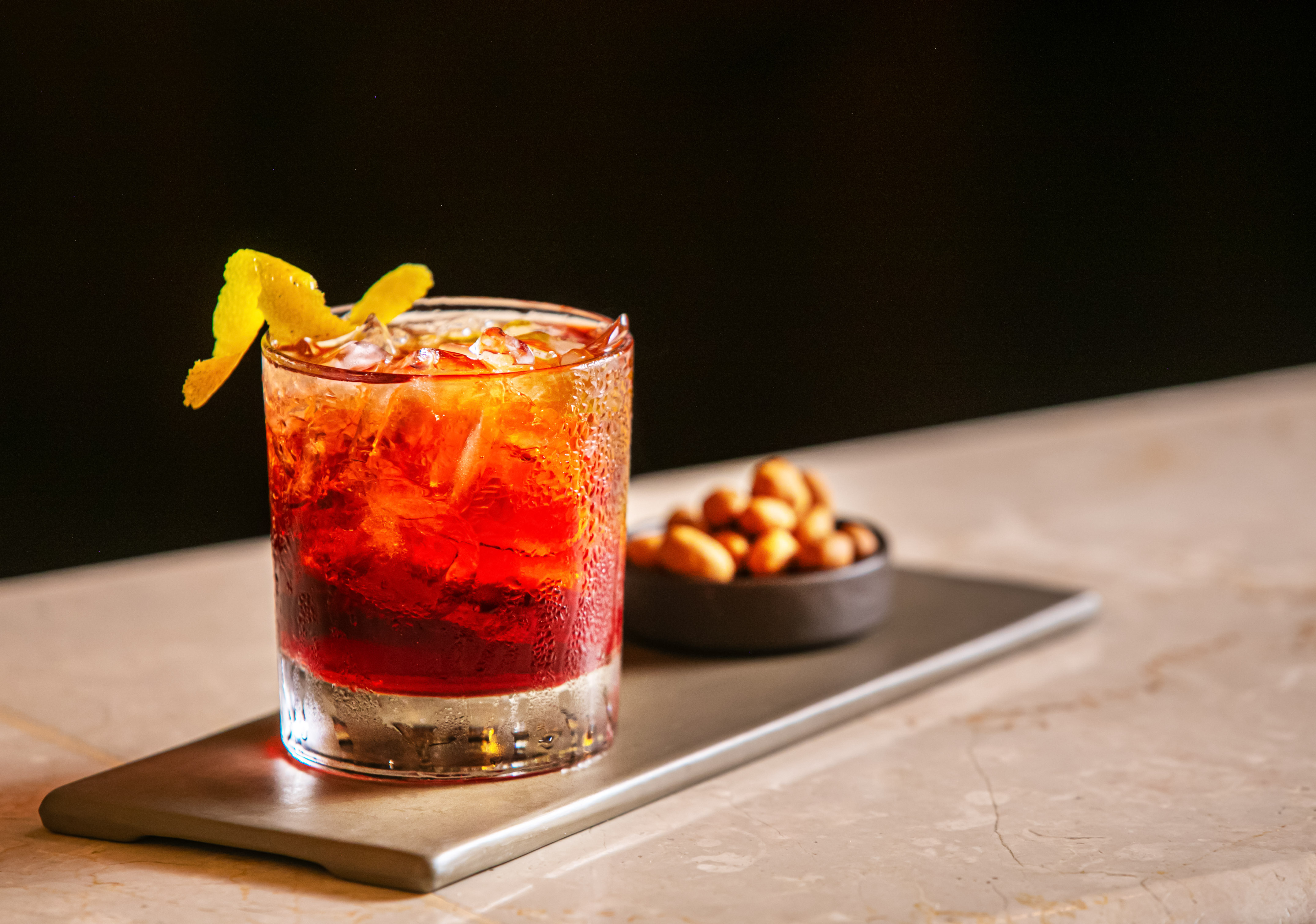 Red cocktail with lemon peel, ice, and peanuts on a tray at a bar.