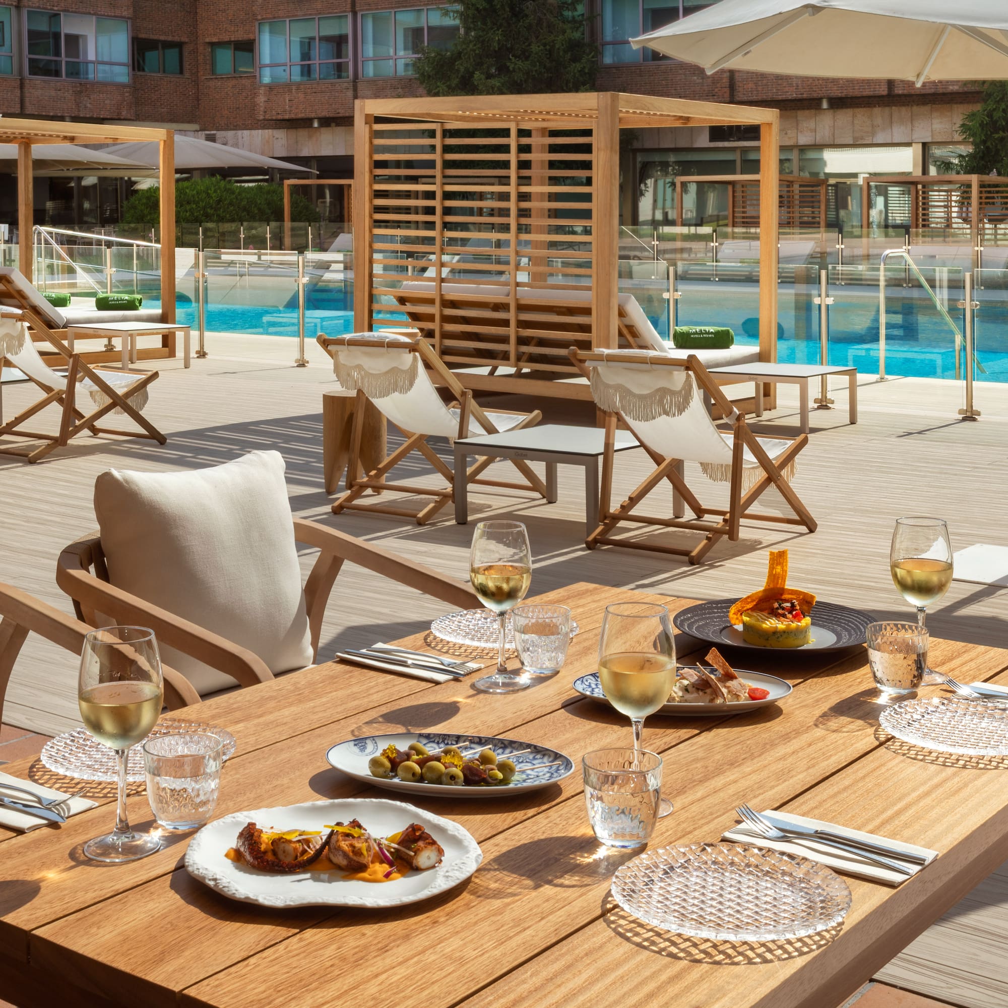 Outdoor dining table with tapas, white wine glasses, by a sunny hotel pool.