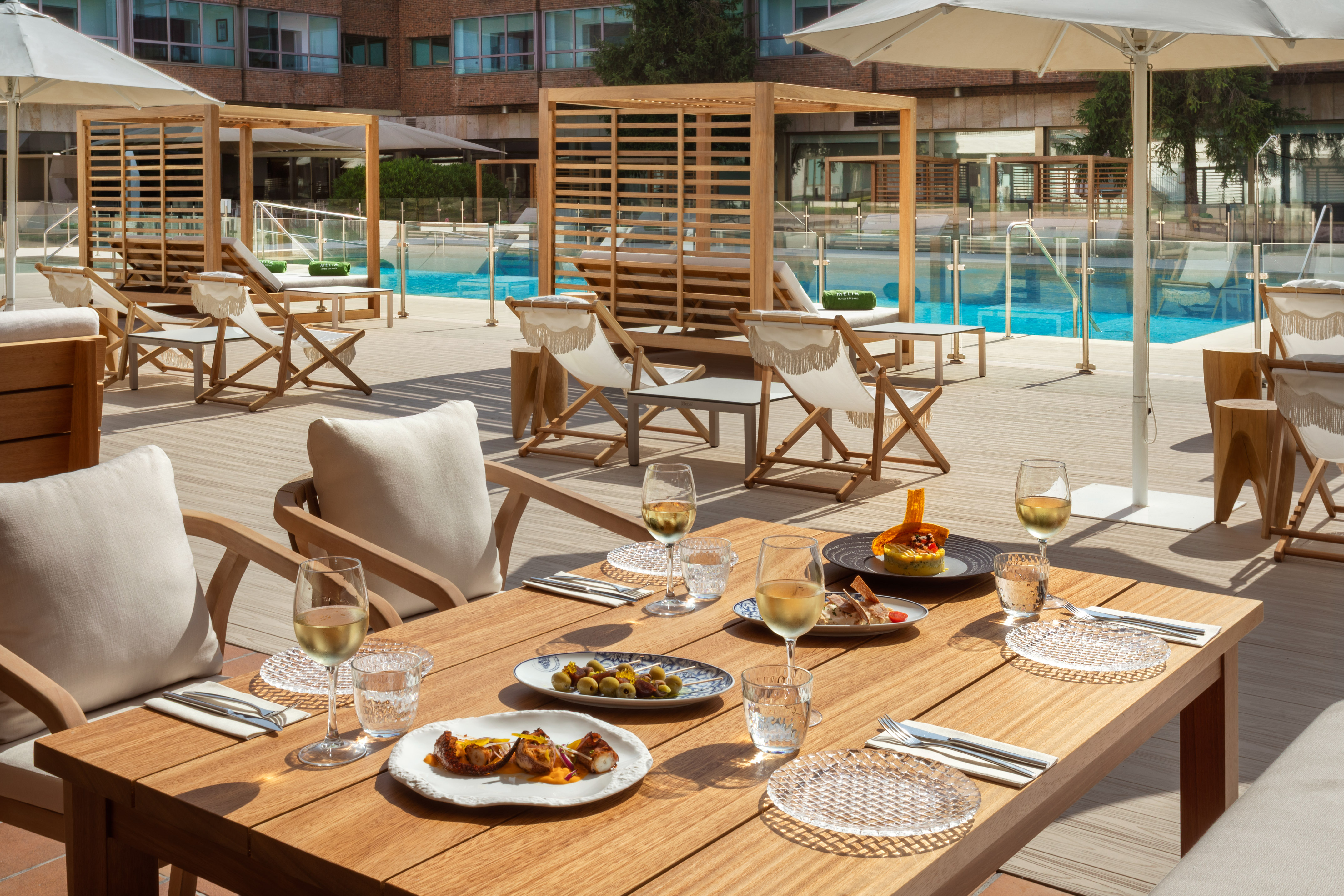 Outdoor dining table with tapas, white wine glasses, by a sunny hotel pool.