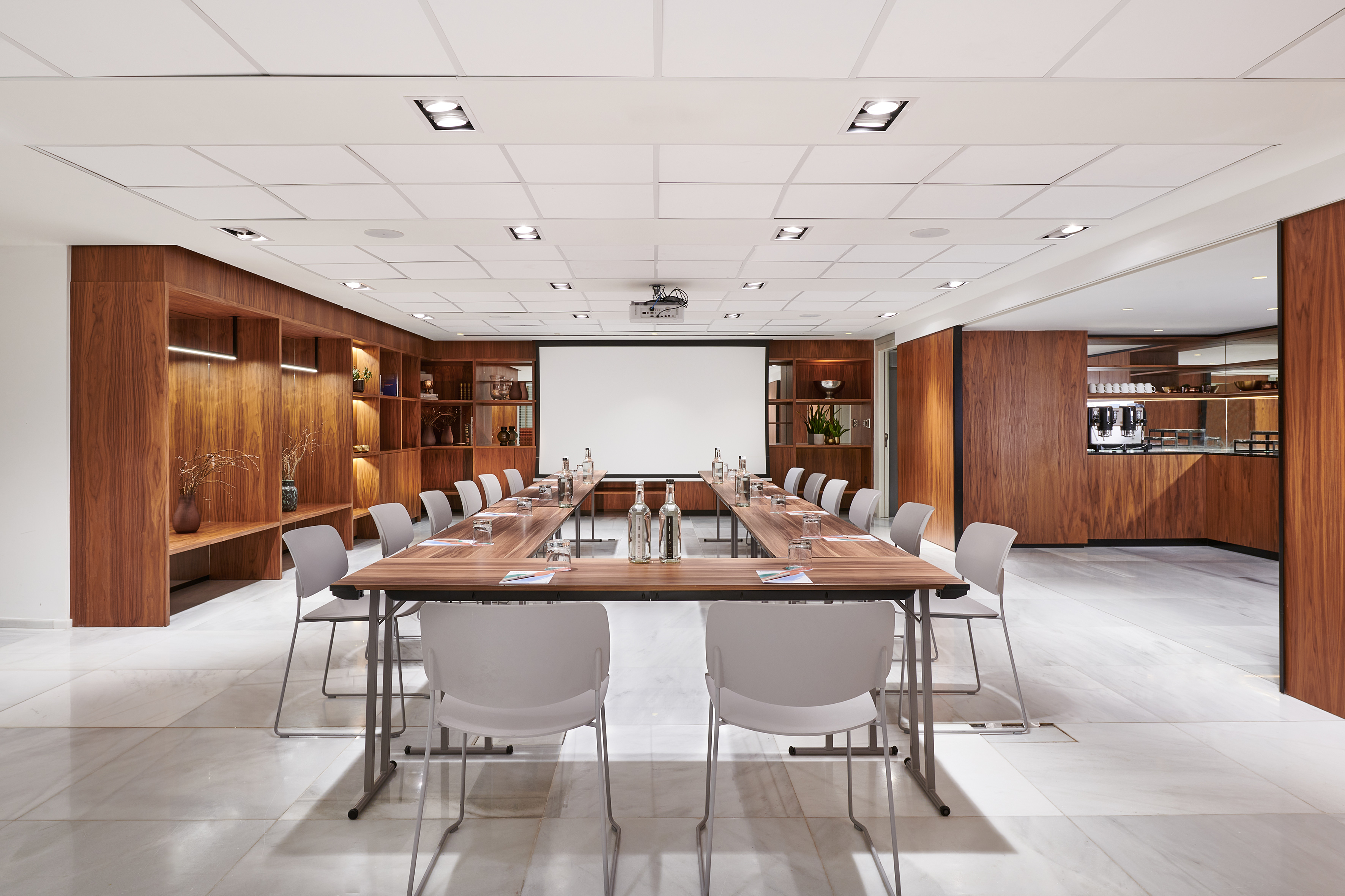 Conference room with U-shaped table, projector screen, wood walls, coffee bar.