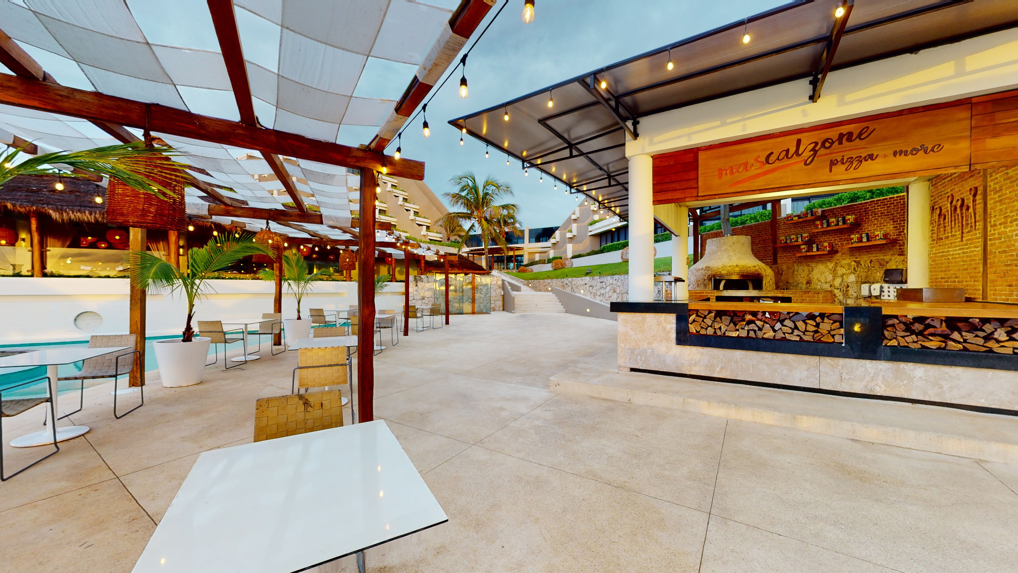 Outdoor resort pizzeria with wood-fired oven, dining area, pool, and string lights.