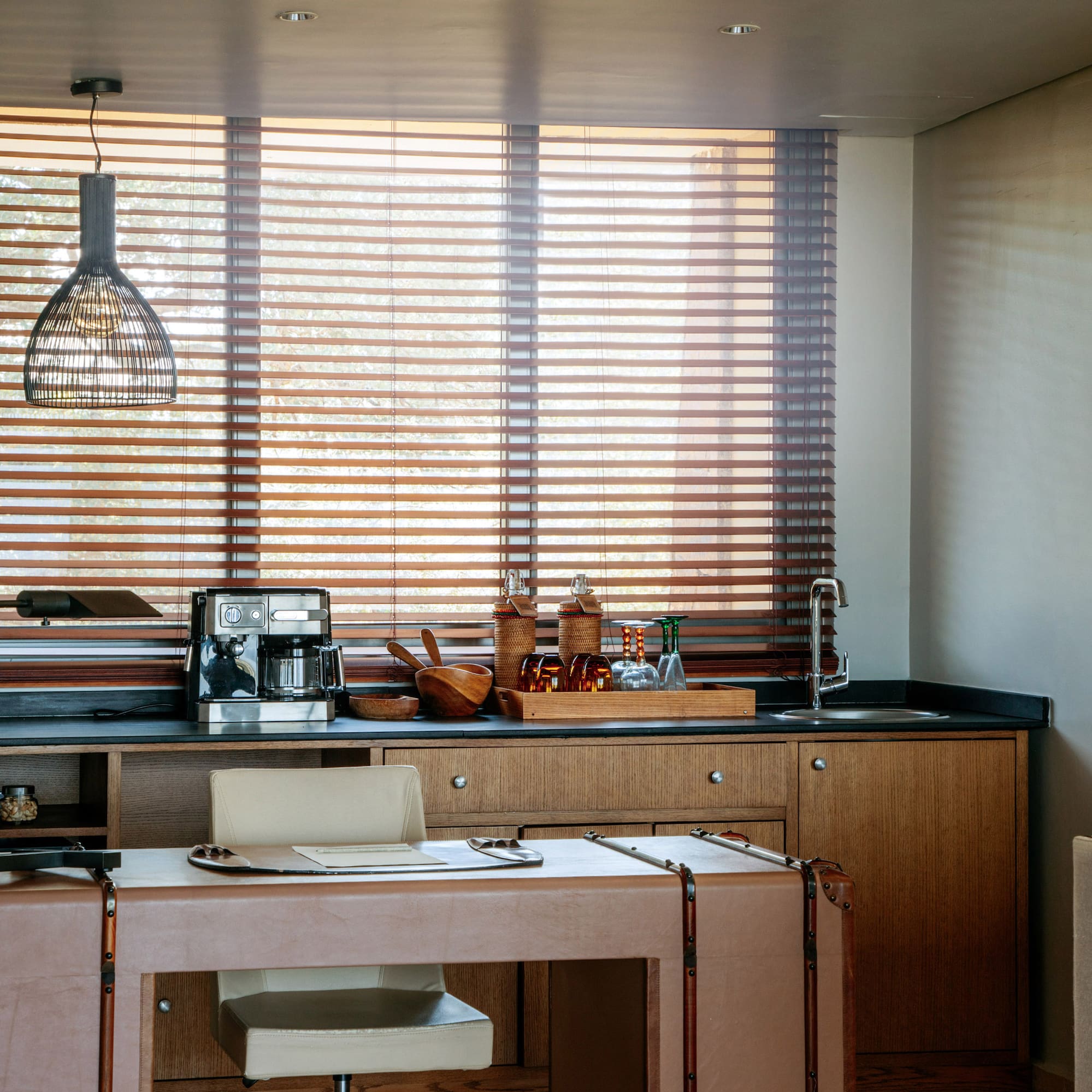 Modern home office with wet bar, coffee machine, leather desk, and wood blinds.