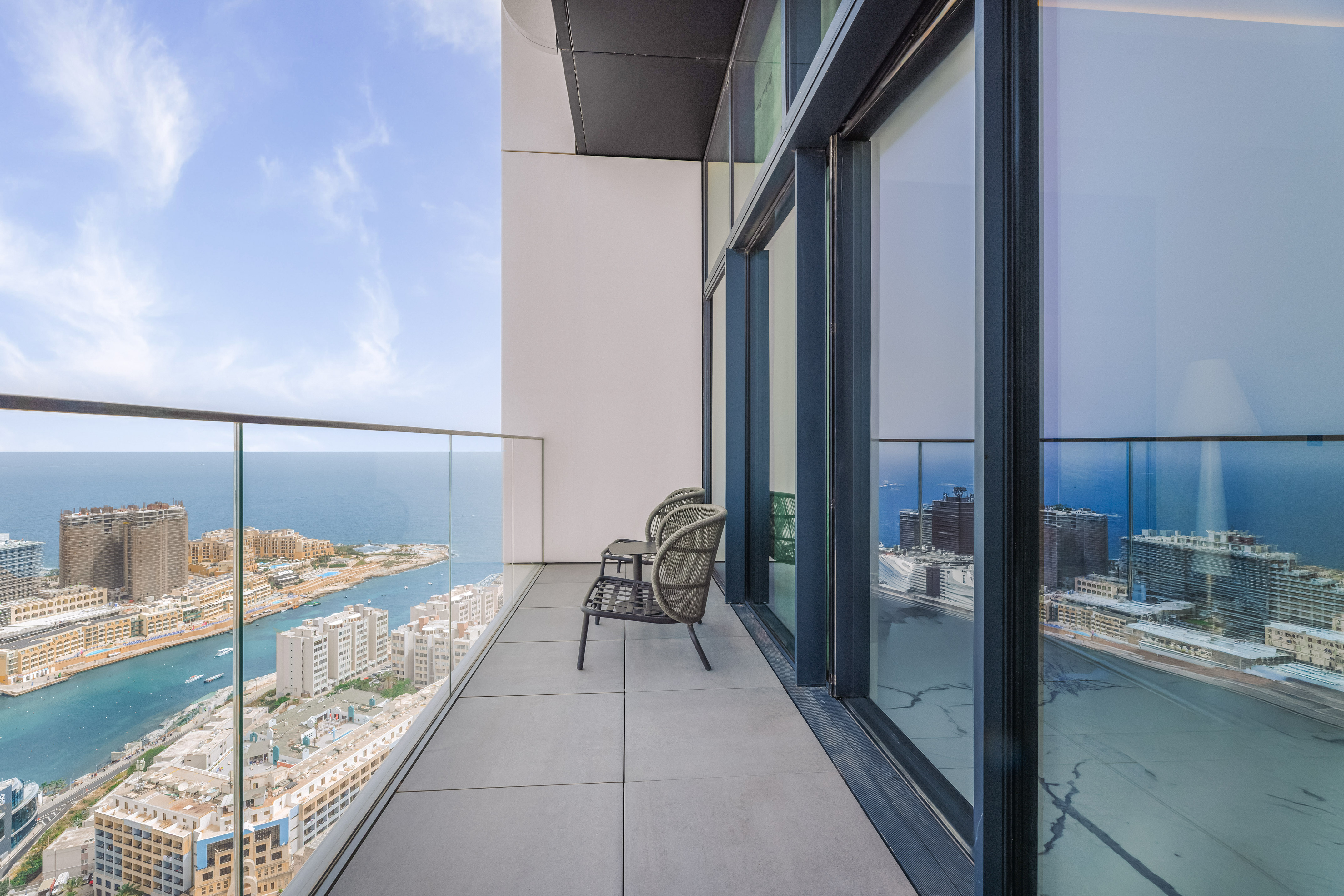 High-rise balcony overlooking sea and coastal city, with outdoor chairs.
