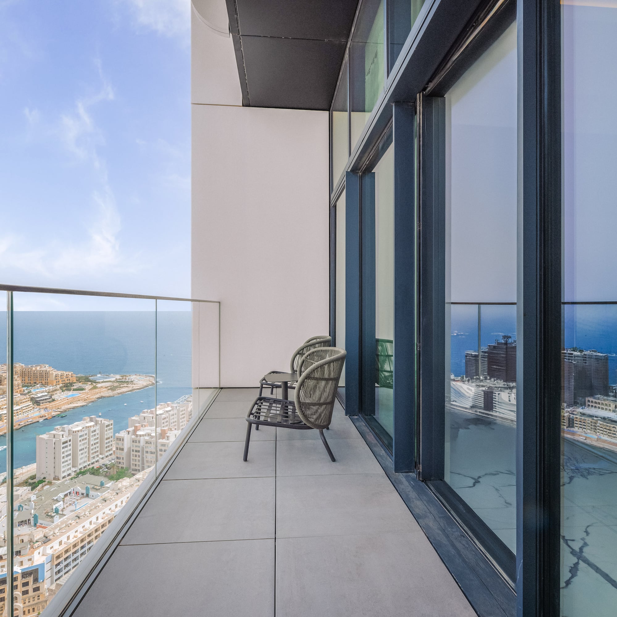 High-rise balcony overlooking sea and coastal city, with outdoor chairs.