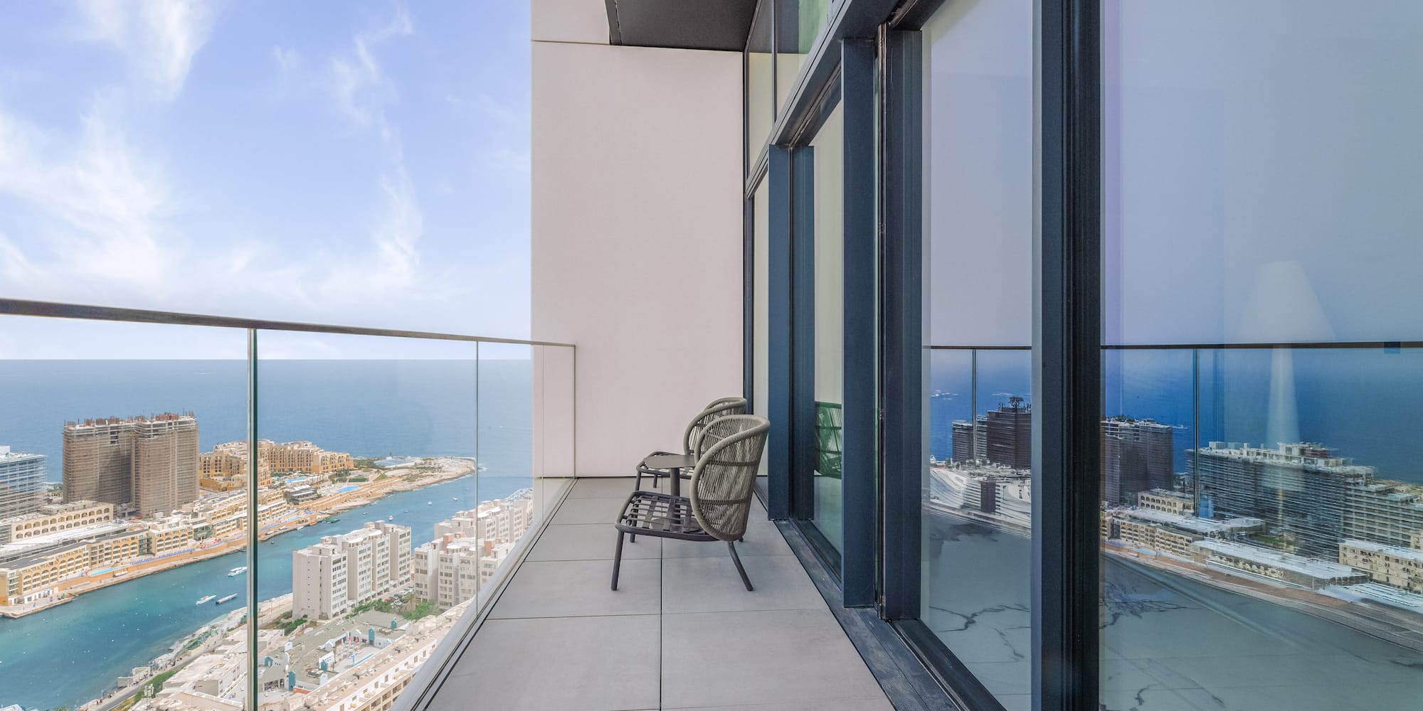 High-rise balcony overlooking sea and coastal city, with outdoor chairs.