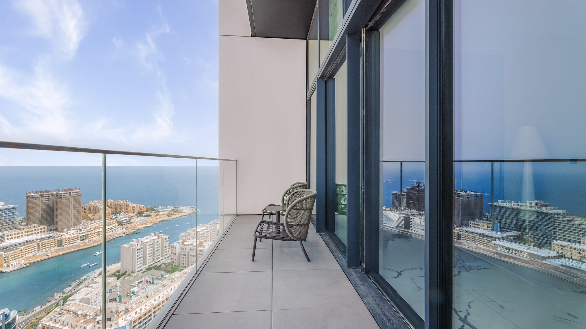 High-rise balcony overlooking sea and coastal city, with outdoor chairs.