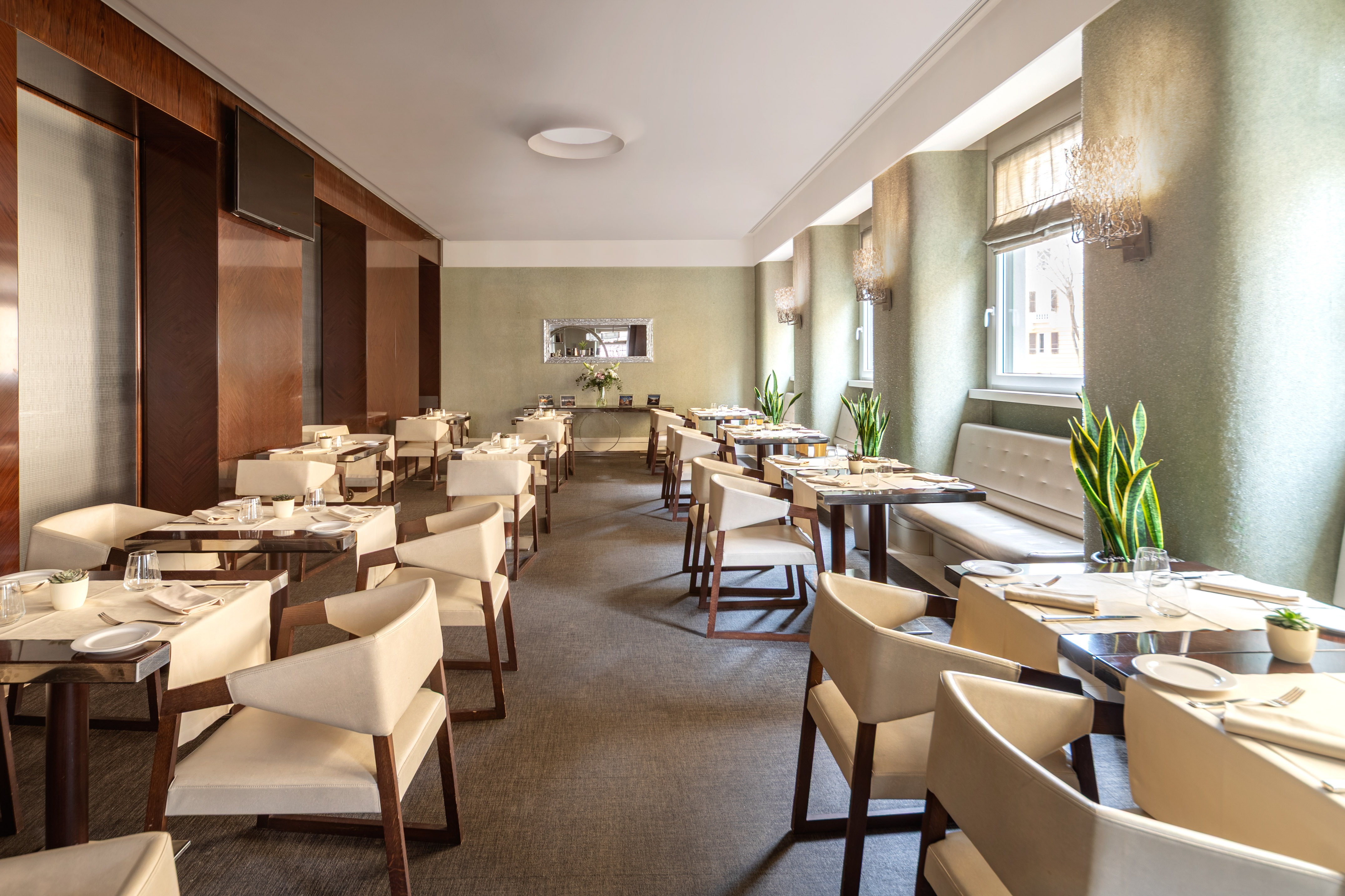 Modern restaurant interior with set dining tables, chairs, banquette, and plants.