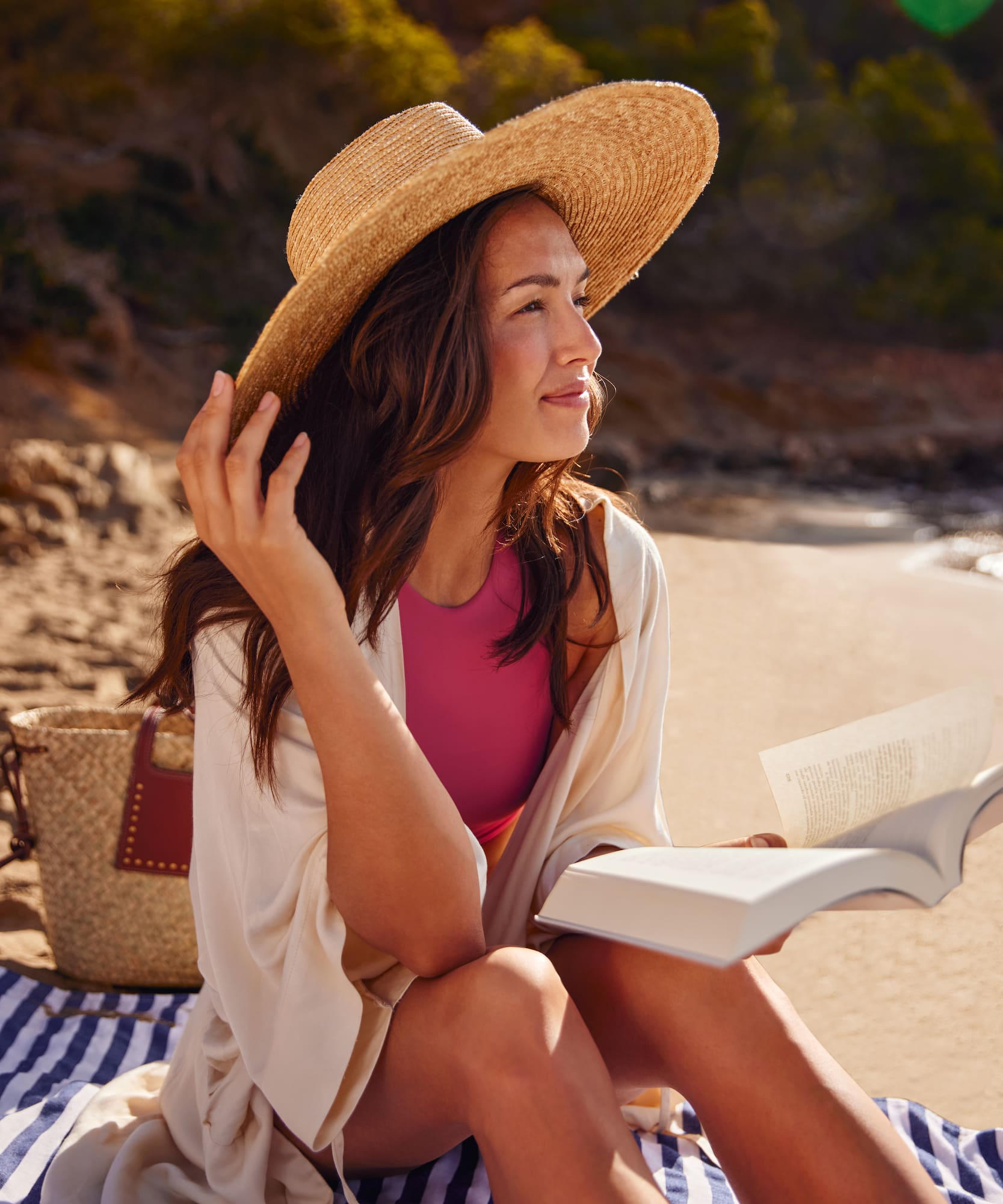 Woman in straw hat, swimsuit, reading book on striped beach blanket.