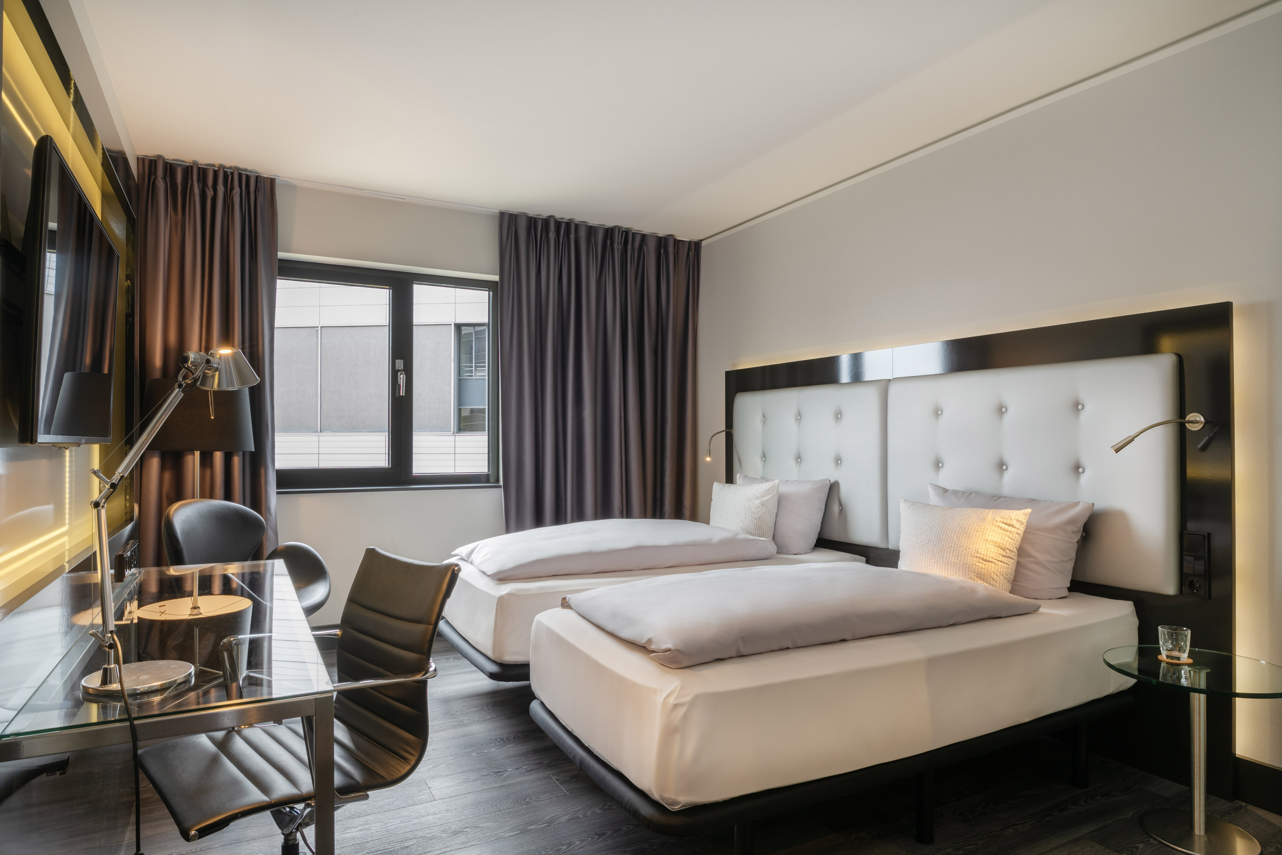 Modern hotel room with two twin beds, work desk, and dark curtains.