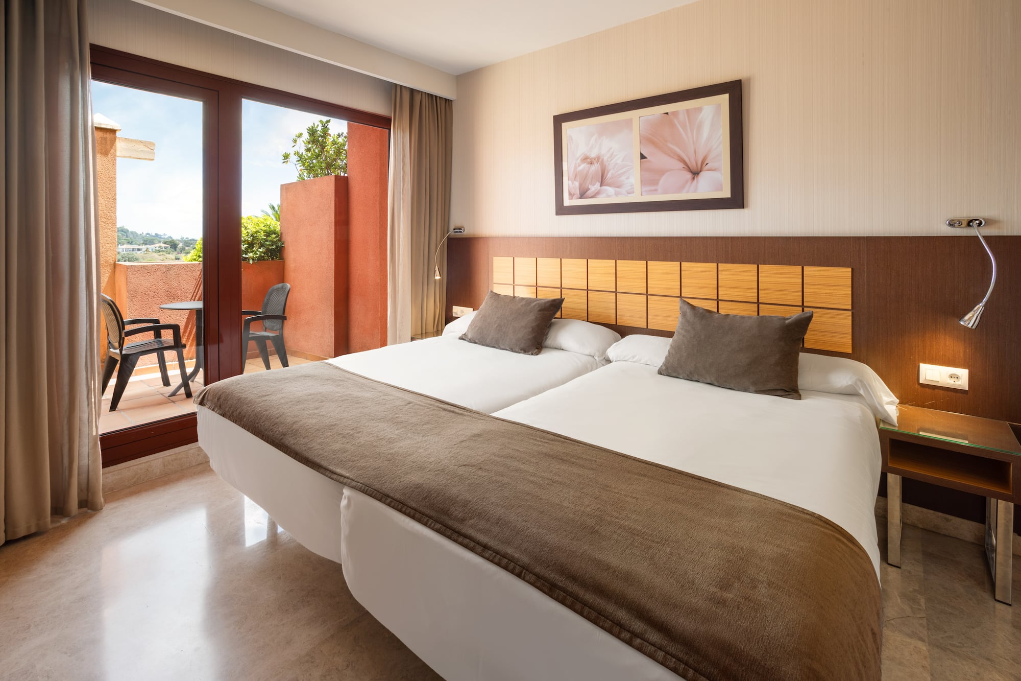 Hotel room with twin beds, wooden headboard, and a balcony with outdoor seating.