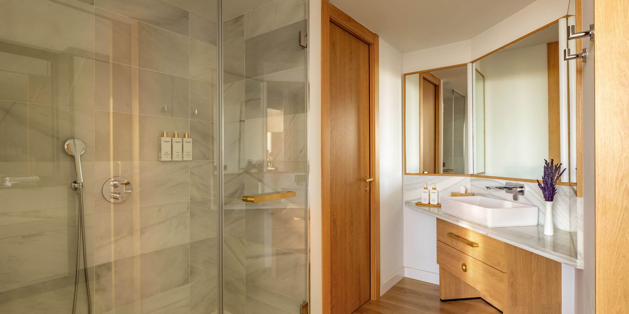 Modern bathroom with glass shower, marble tiles, wood vanity, and gold accents.