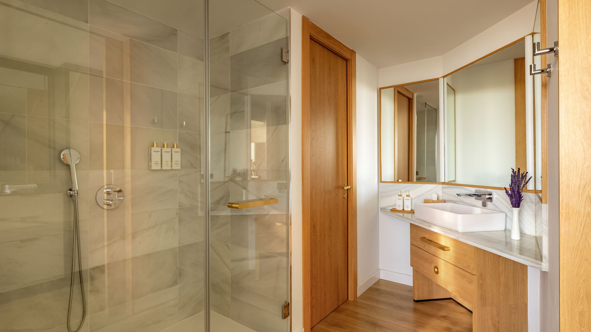 Modern bathroom with glass shower, marble tiles, wood vanity, and gold accents.