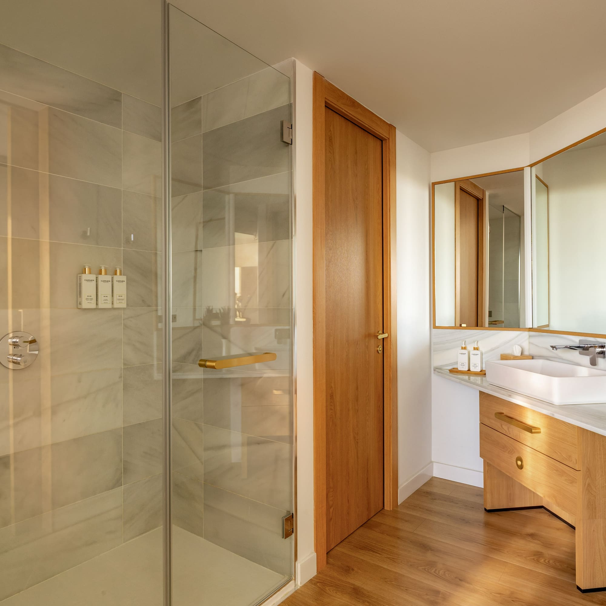 Modern bathroom with glass shower, marble tiles, wood vanity, and gold accents.