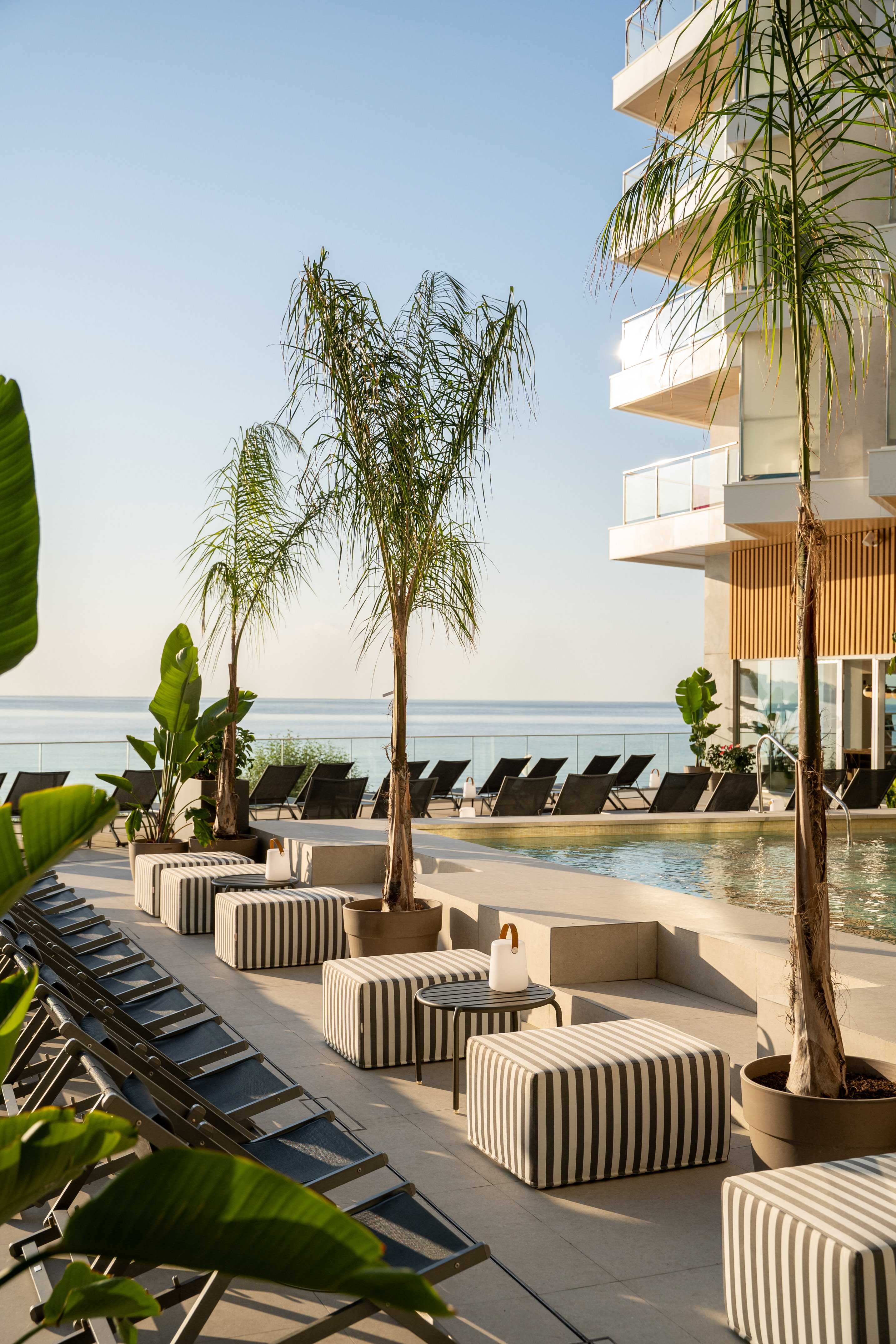 Resort pool deck with ocean view, sunbeds, striped ottomans, palm trees.