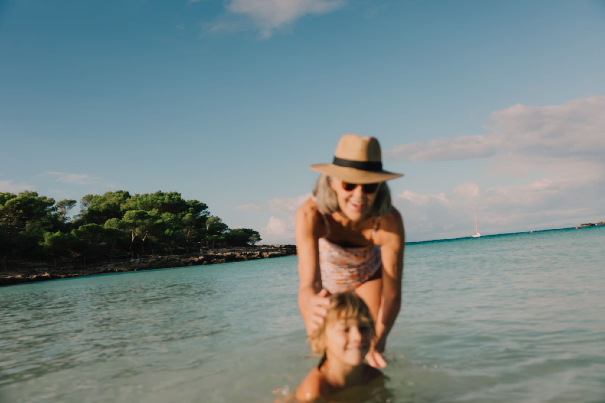 Abuela y nieta sonriendo jugando en el mar, playa rocosa y cielo azul.
