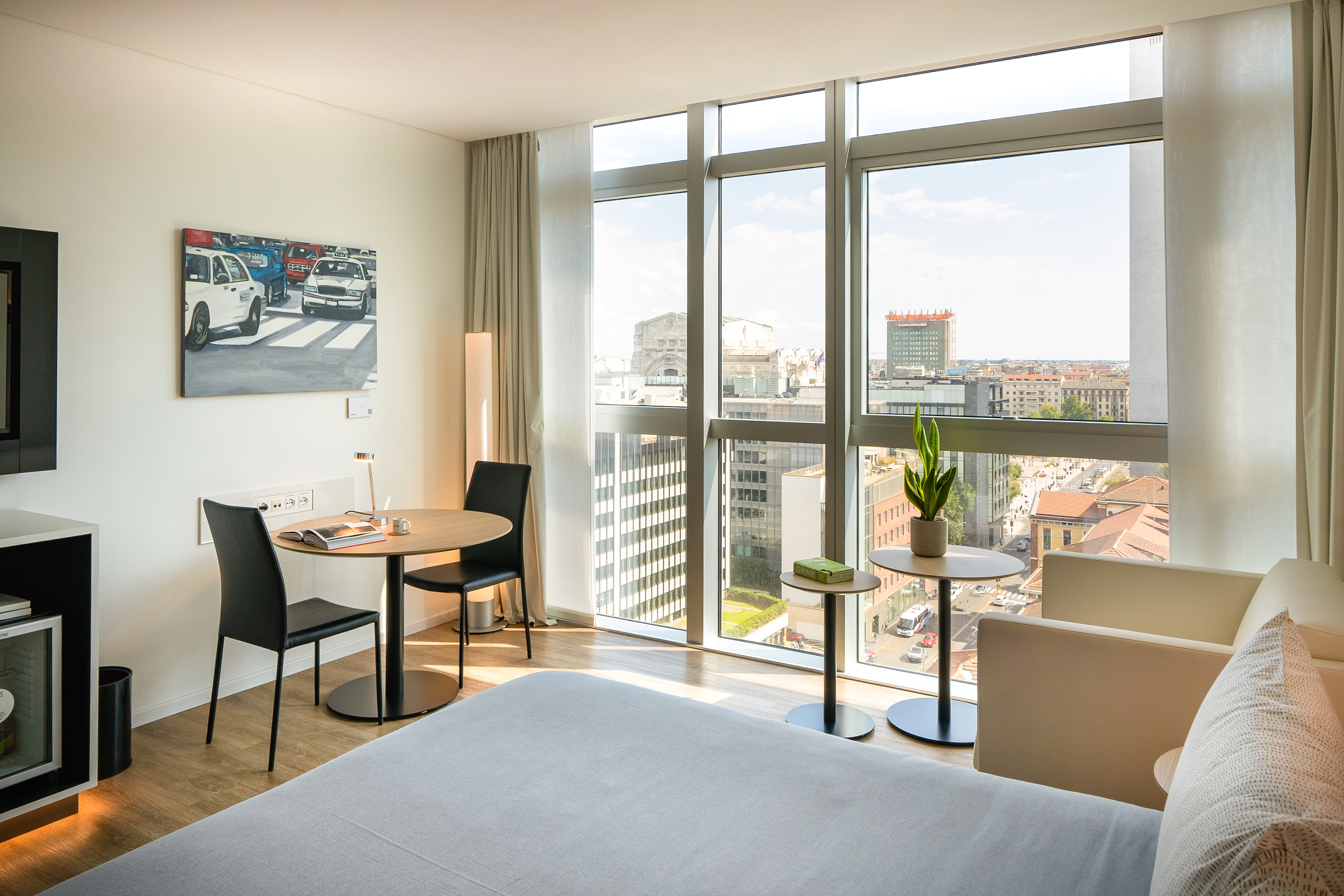 Modern hotel room with city view, large window, bed, and dining area.
