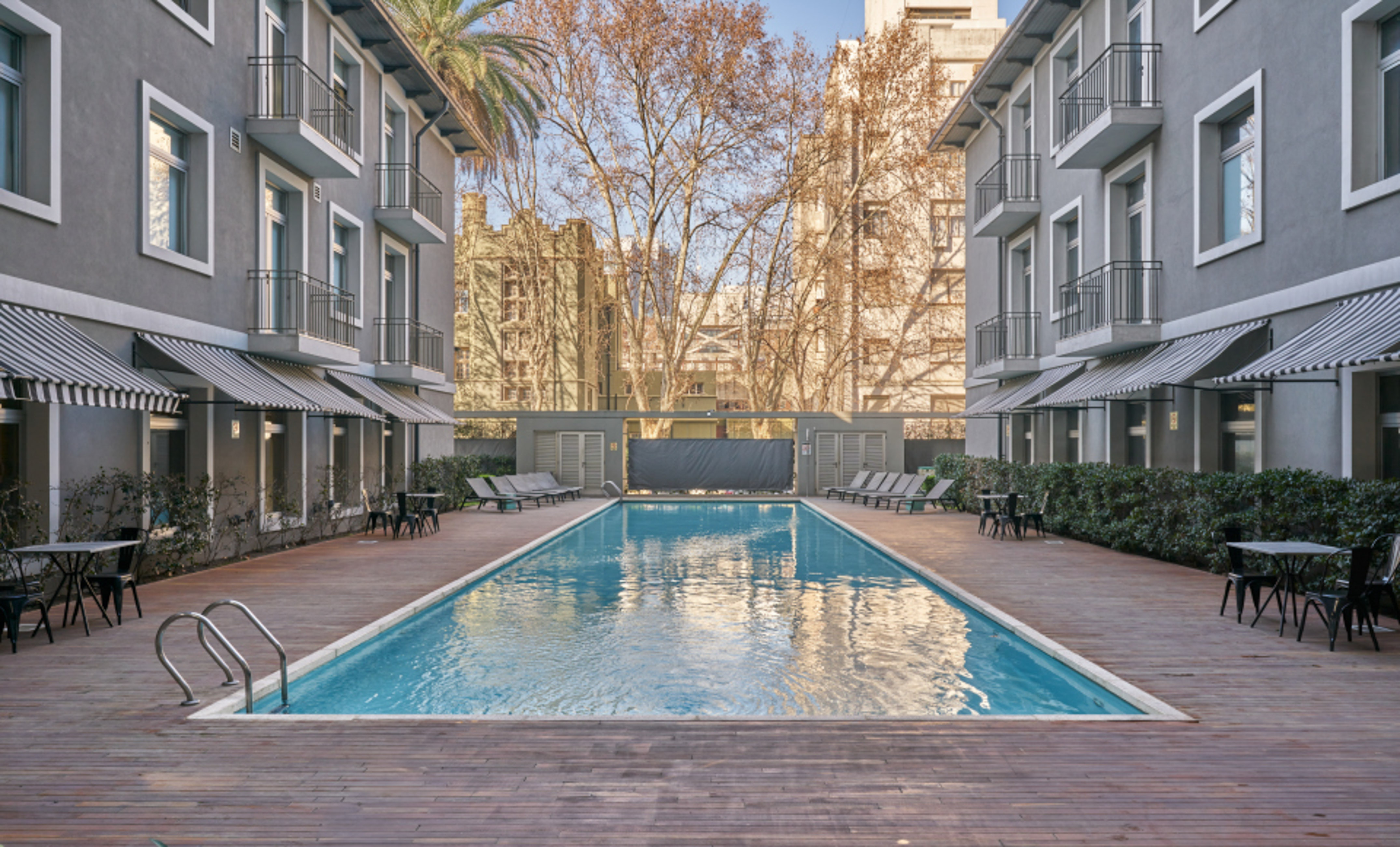 Outdoor swimming pool, wooden deck, hotel buildings with striped awnings.