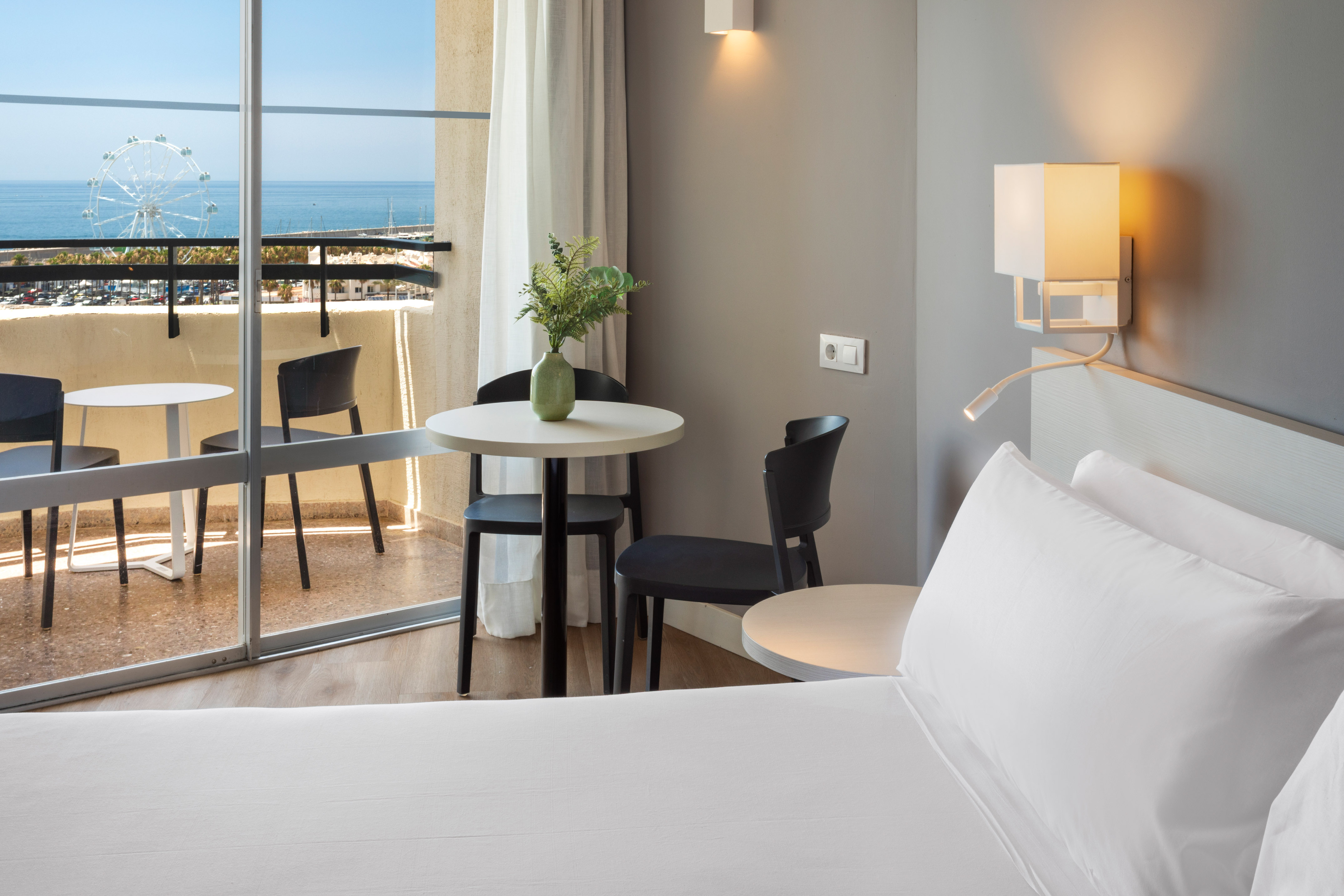 Modern hotel room with sea view balcony, distant Ferris wheel, and comfortable bed.