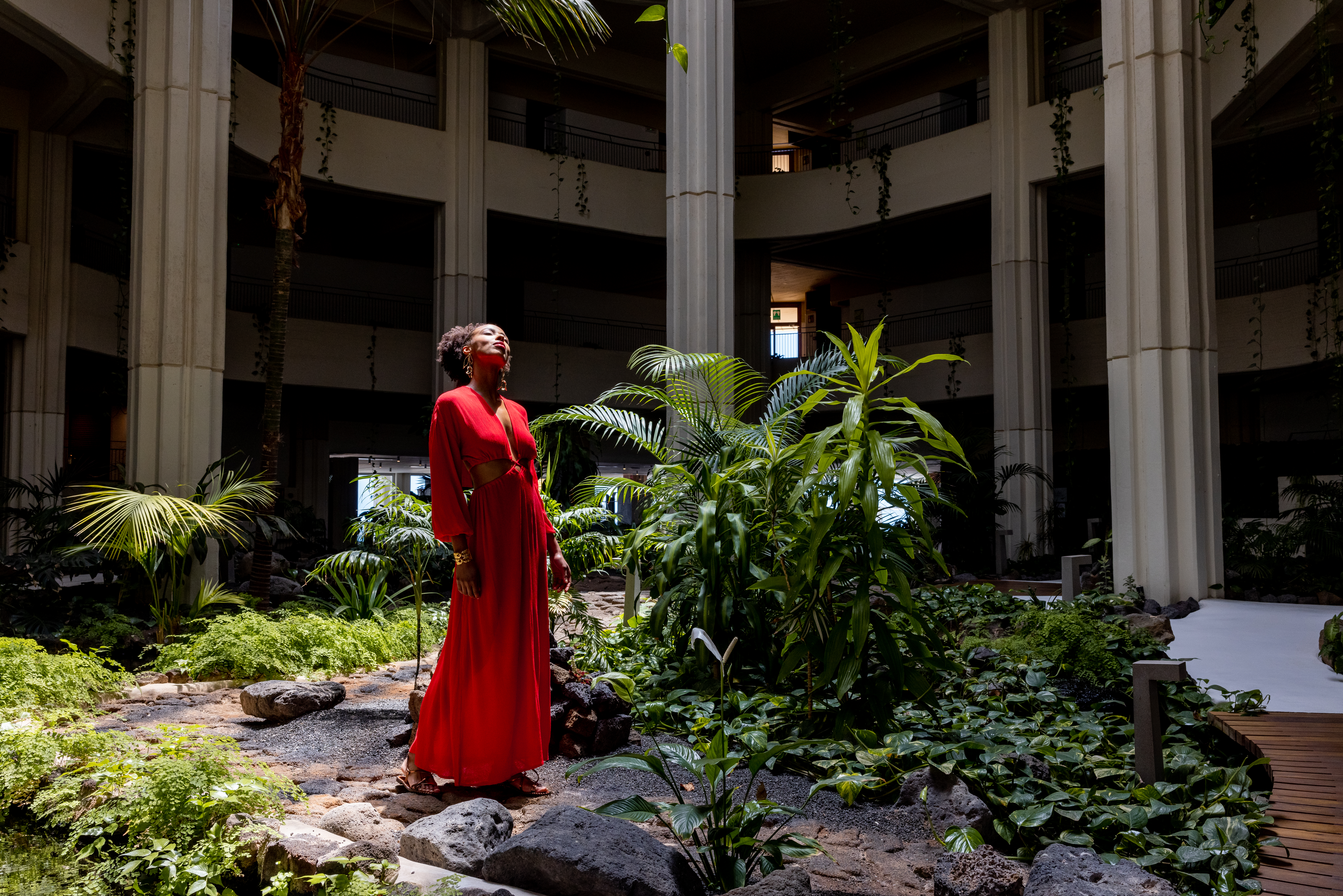 Woman in red dress in lush tropical indoor garden atrium, looking up.