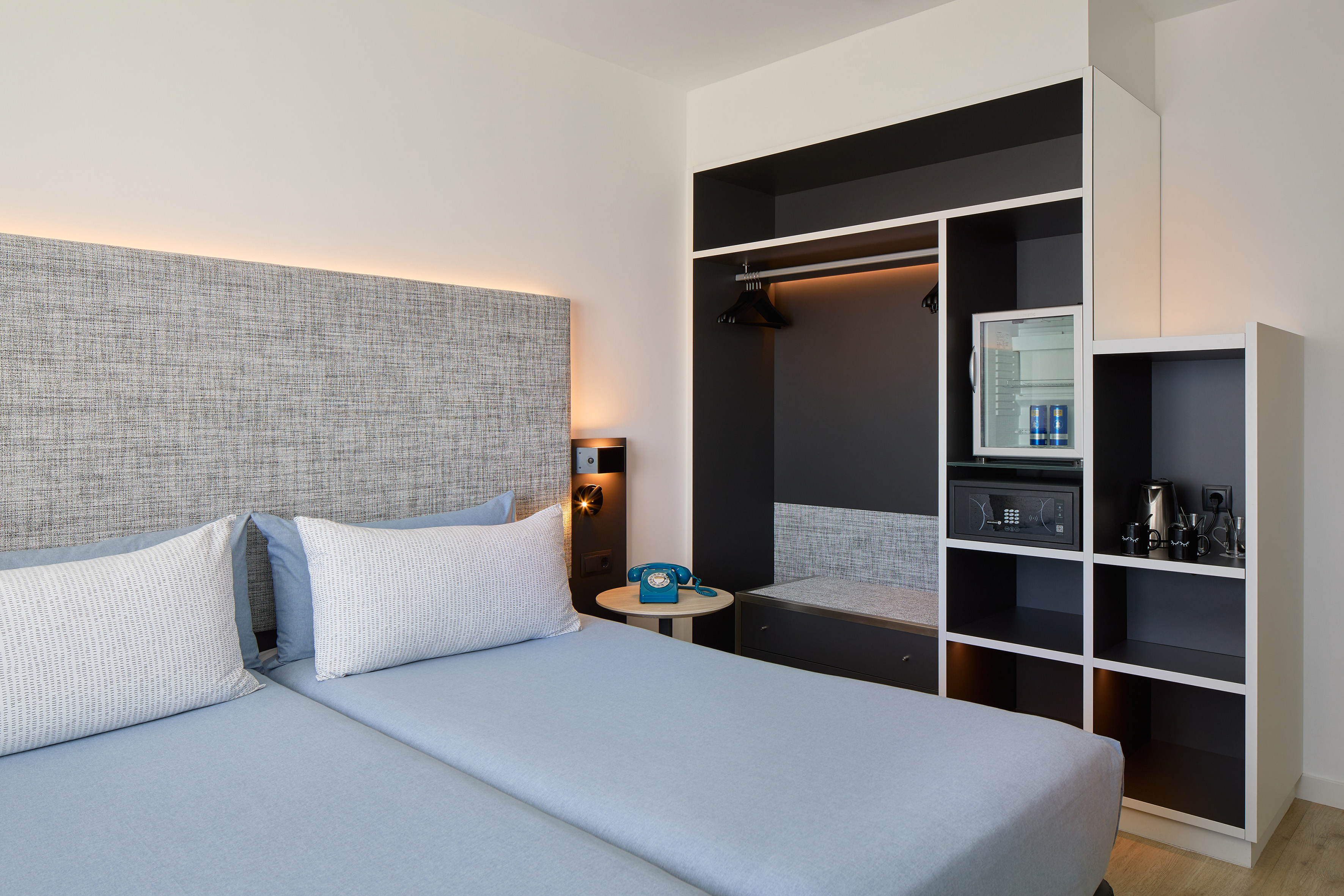 Modern hotel room with two beds, blue bedding, wardrobe, mini-fridge, and safe.