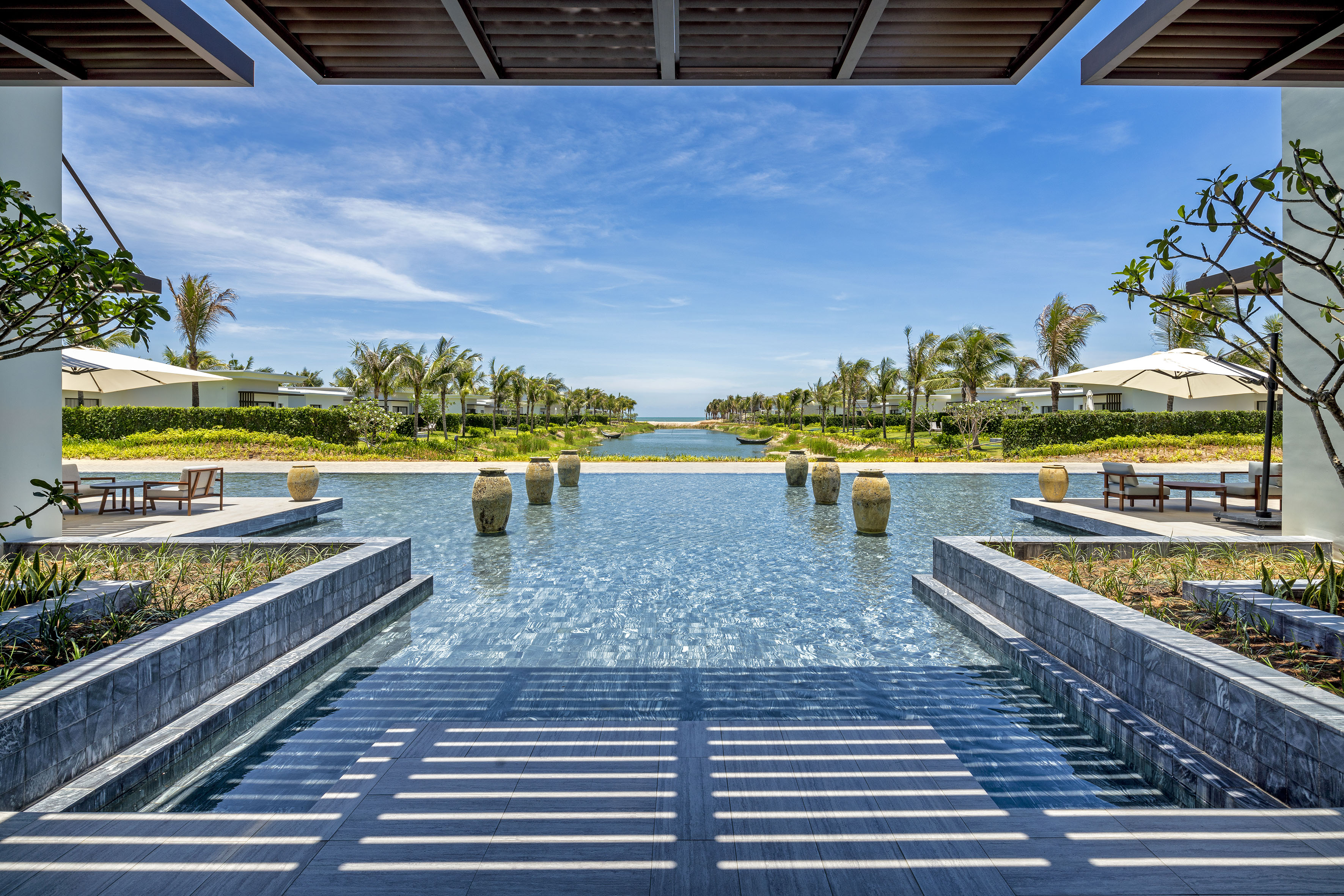 Tropical resort pool leading to ocean canal, flanked by palms and villas.