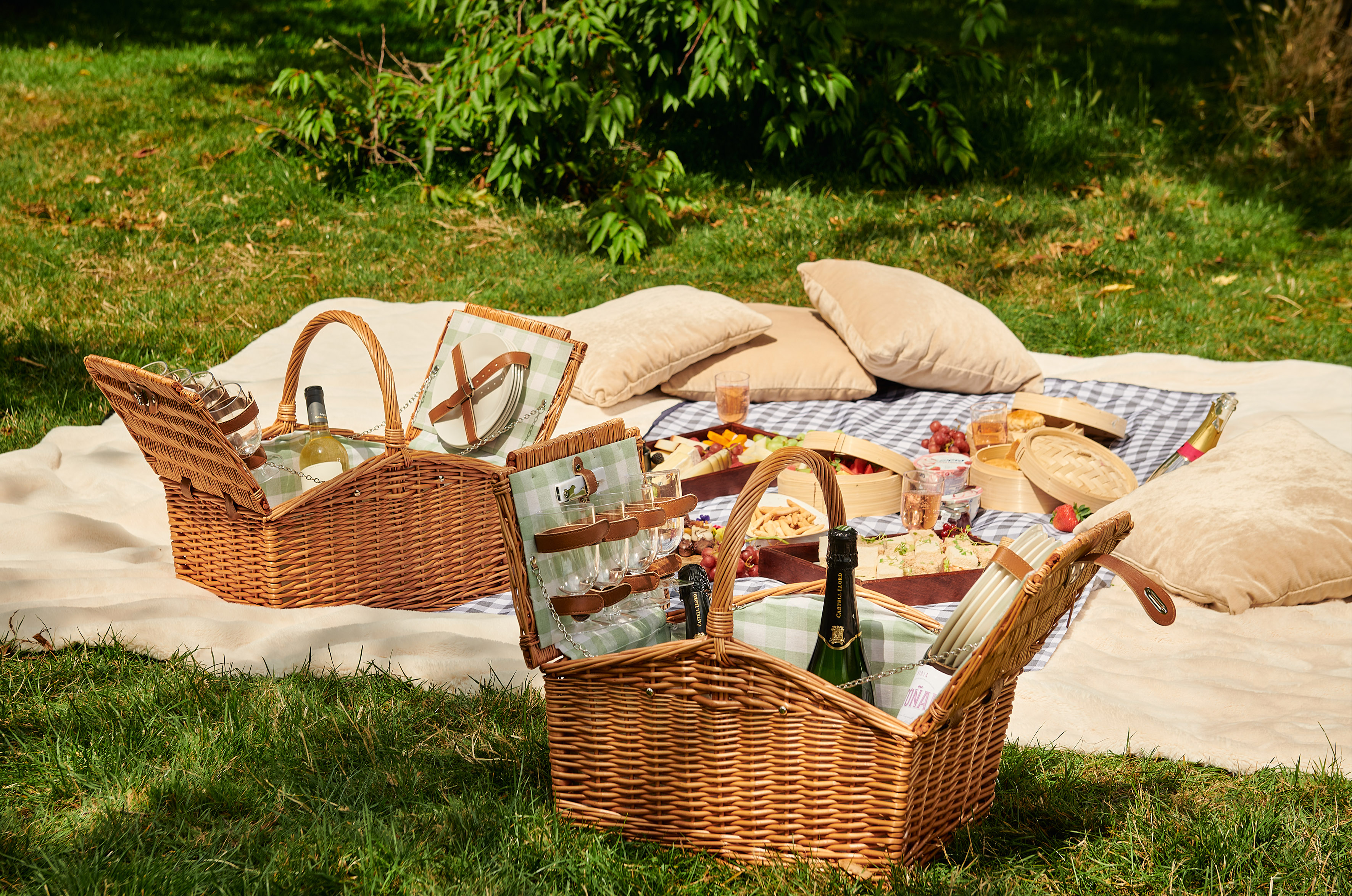 Outdoor picnic with two wicker baskets, food, wine, and blankets on green grass.