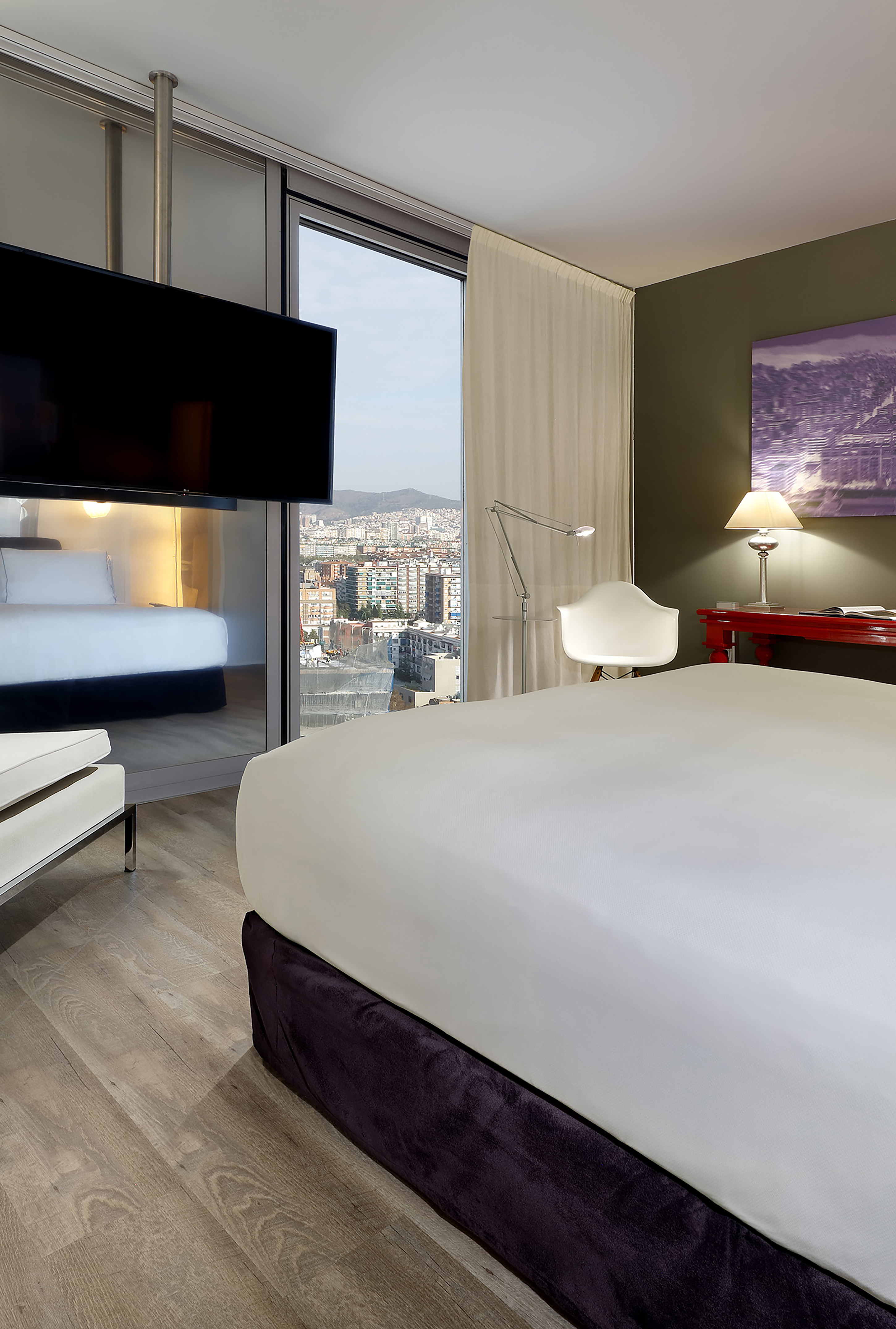 Modern hotel room: city view window, comfortable bed, red desk, and mounted TV.
